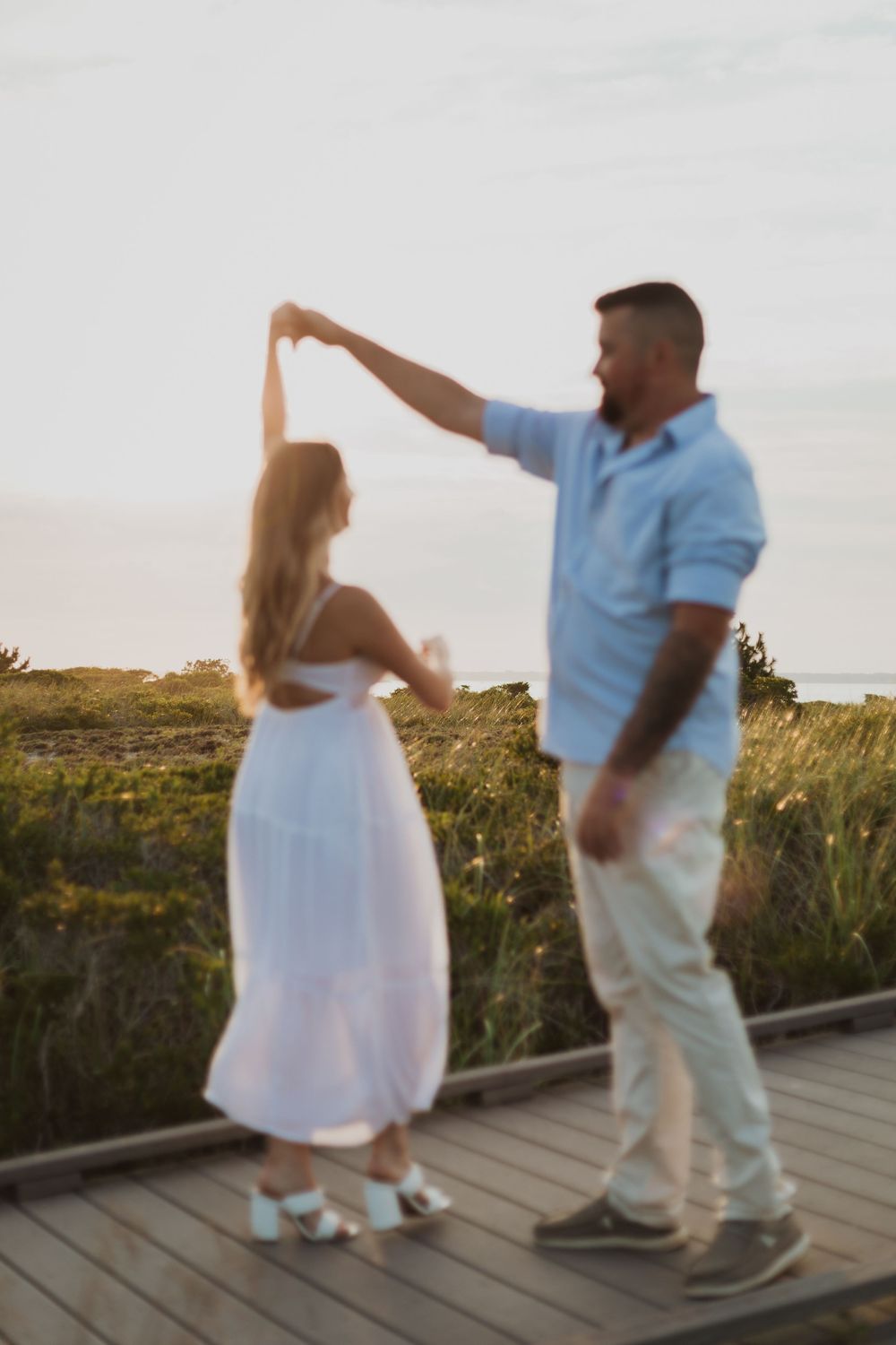 A couple dancing together on a wooden boardwalk at sunset with the sky and grassy field in the background.