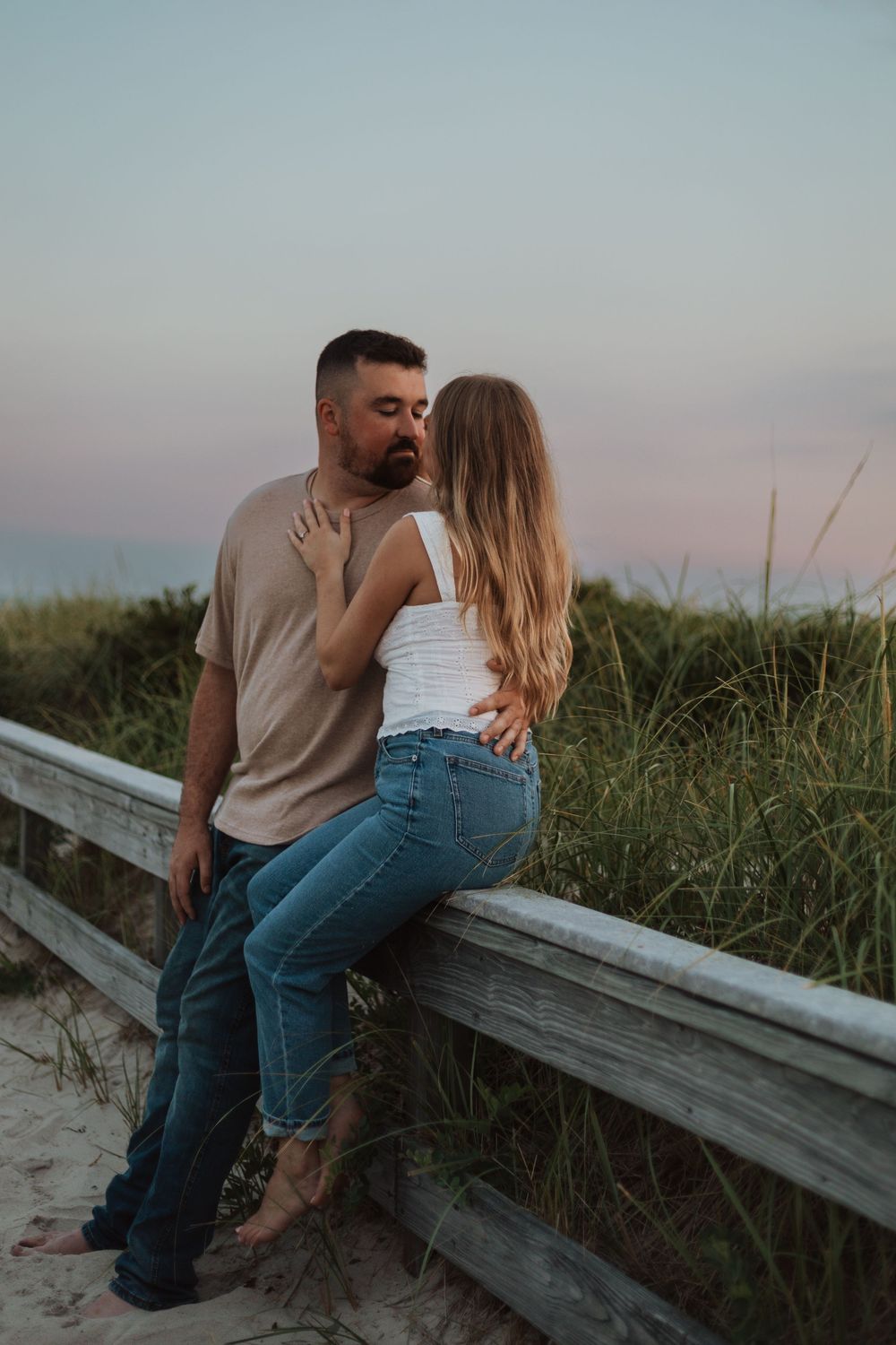 A romantic couple embraces on a wooden boardwalk along a beach at dusk with sea grass in the background.