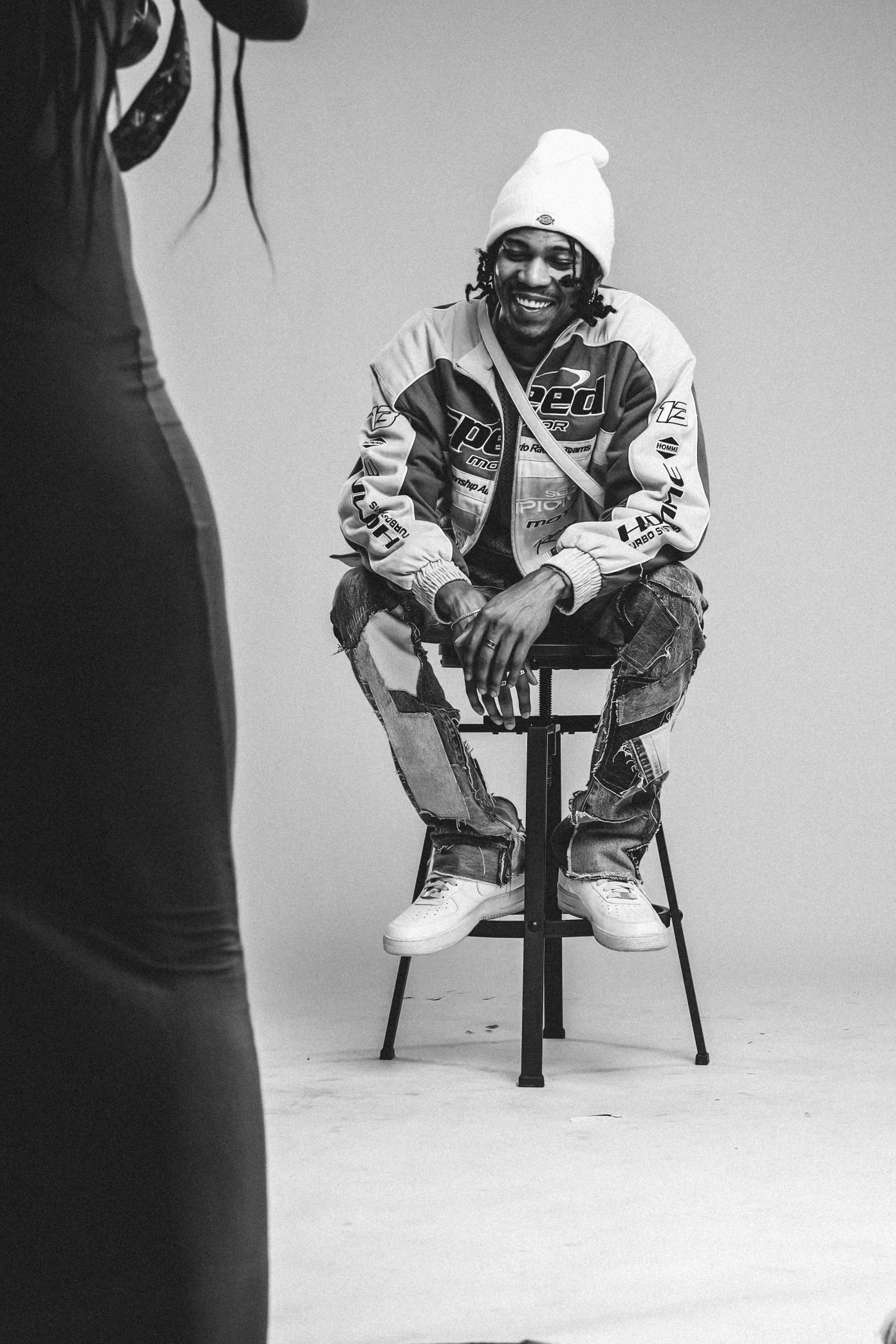 Black and white studio portrait of person in winter attire sitting on stool.