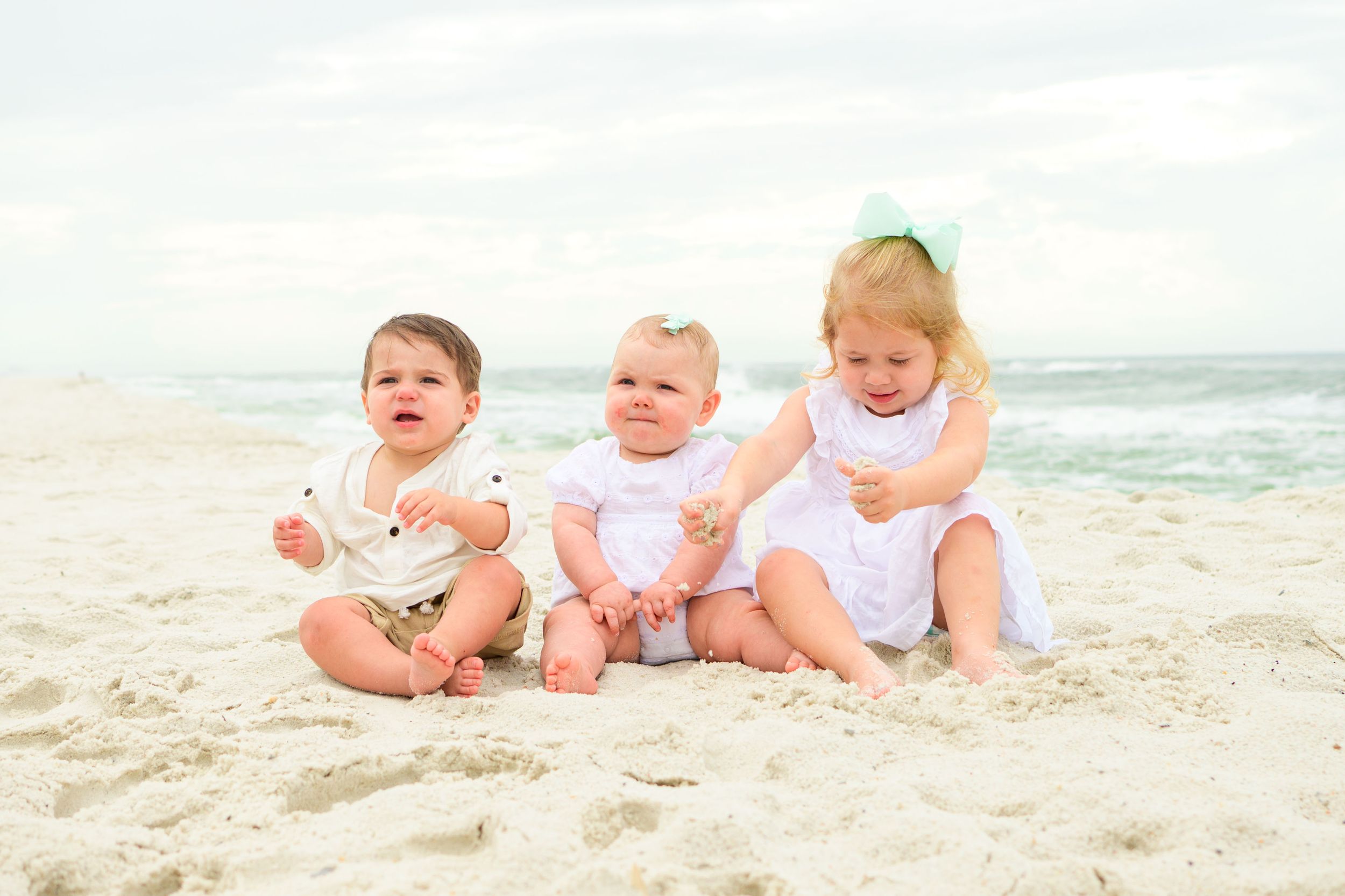Three small children in white outfits sitting together on the sand at the beach.