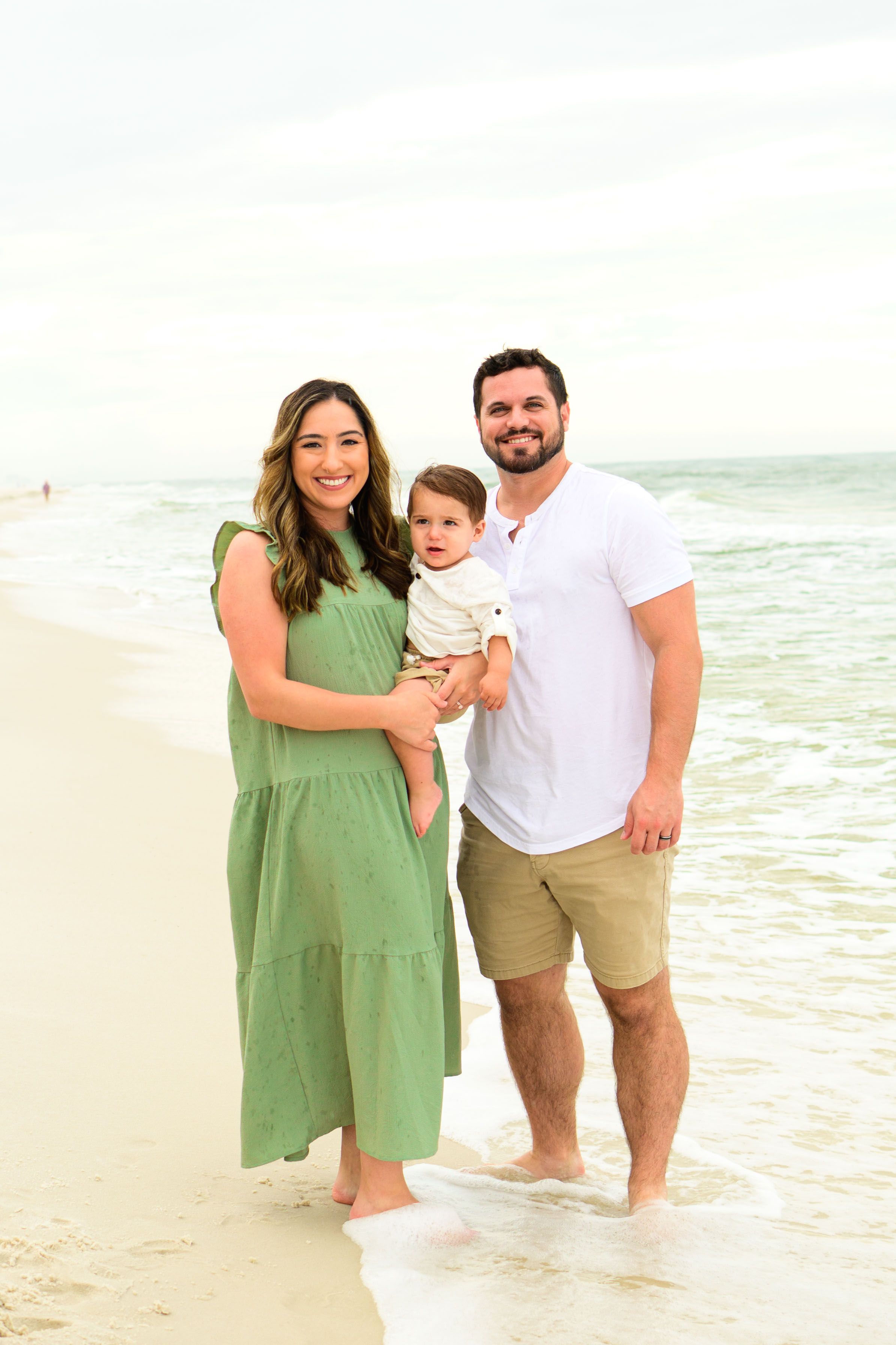 A family in summer attire poses together on a white sandy beach with ocean waves behind them.