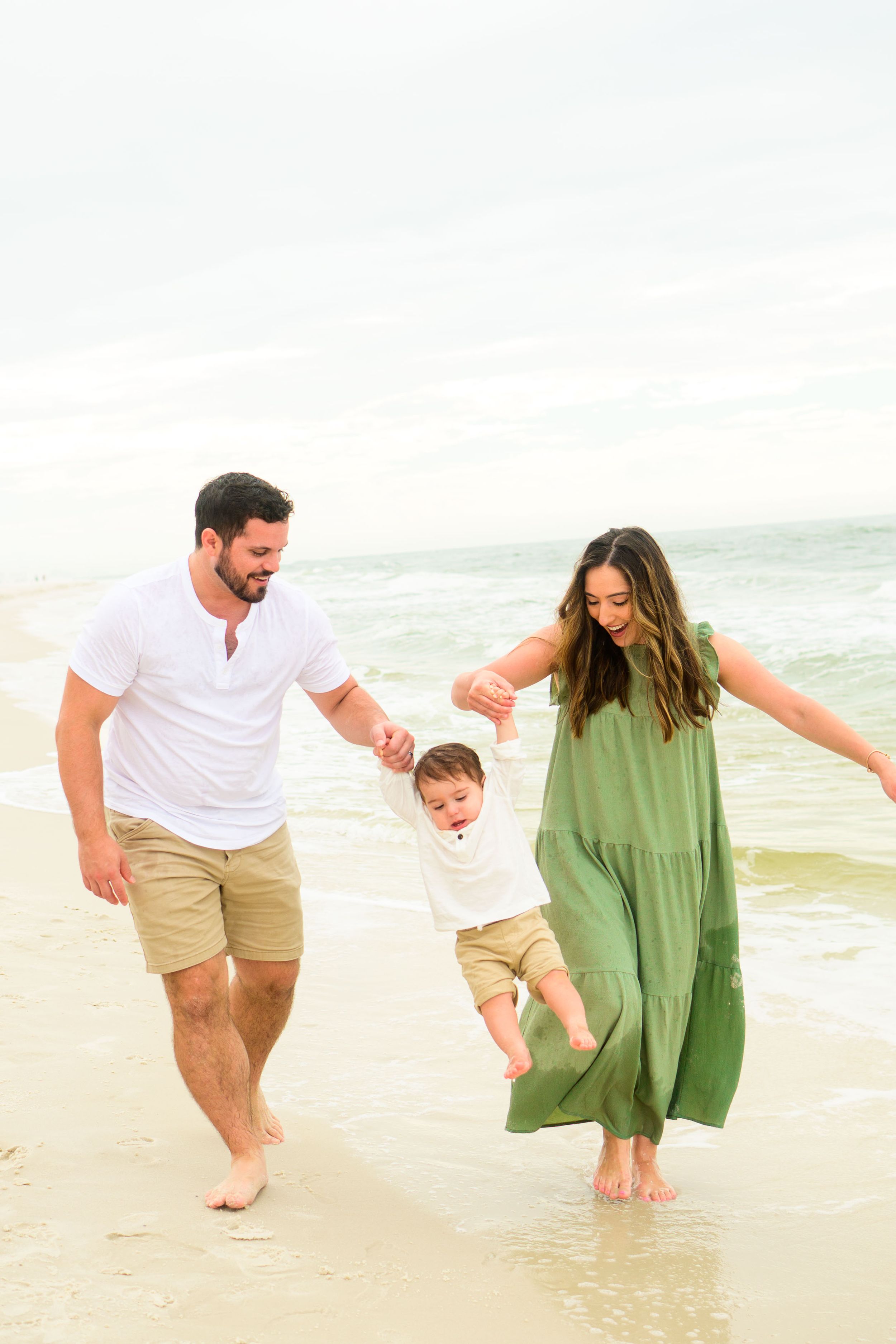 Family swings child while walking together on beach in casual summer attire.
