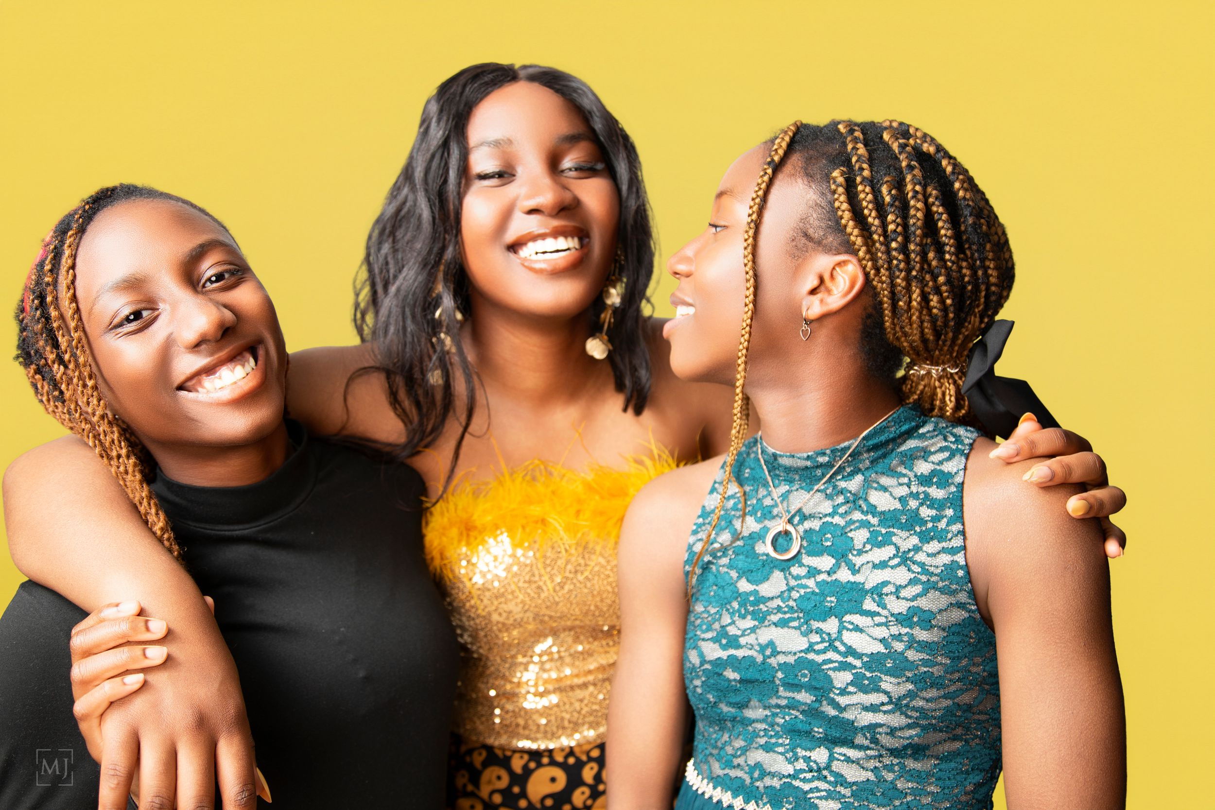 Three sisters share a joyful moment while posing against a bright yellow background.