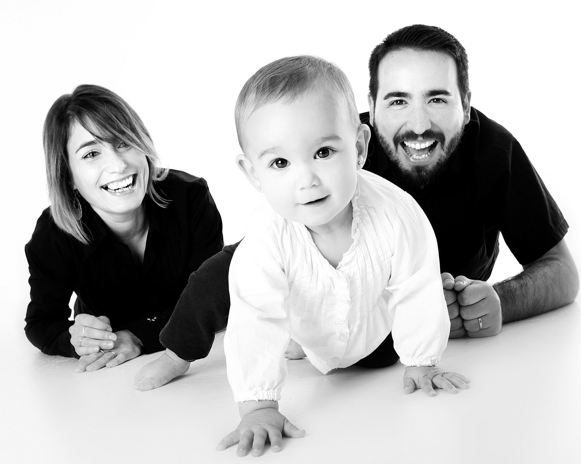 Black and white family portrait with a baby crawling between parents on white background.