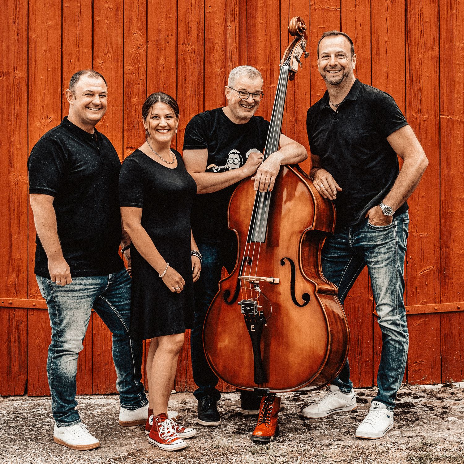 A musical group poses in front of a rustic red barn wall with instruments including an upright bass and guitar.