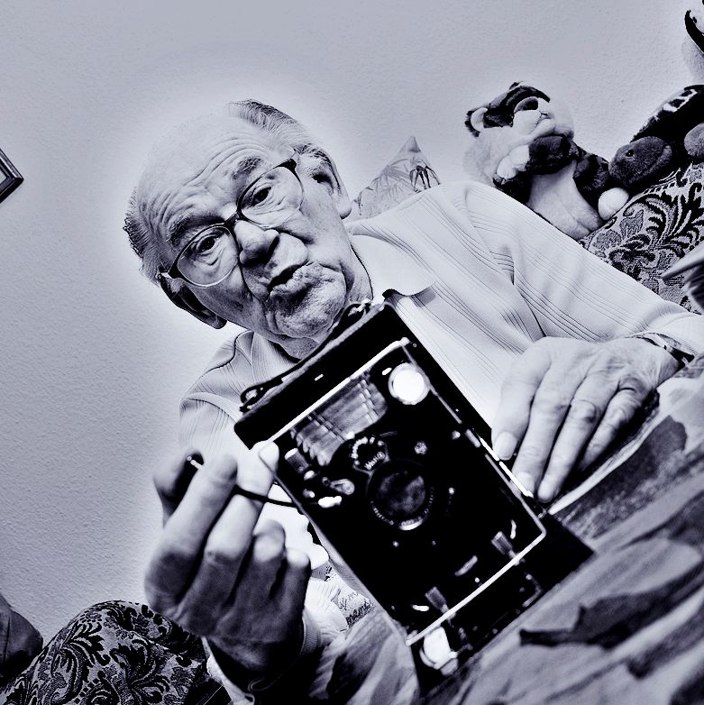 lebensfrohes Portrait eines älteren Mannes mit seinem älteren Fotoapparat auf dem Sofa seines Zimmers im Altersheim in Bad Arolsen