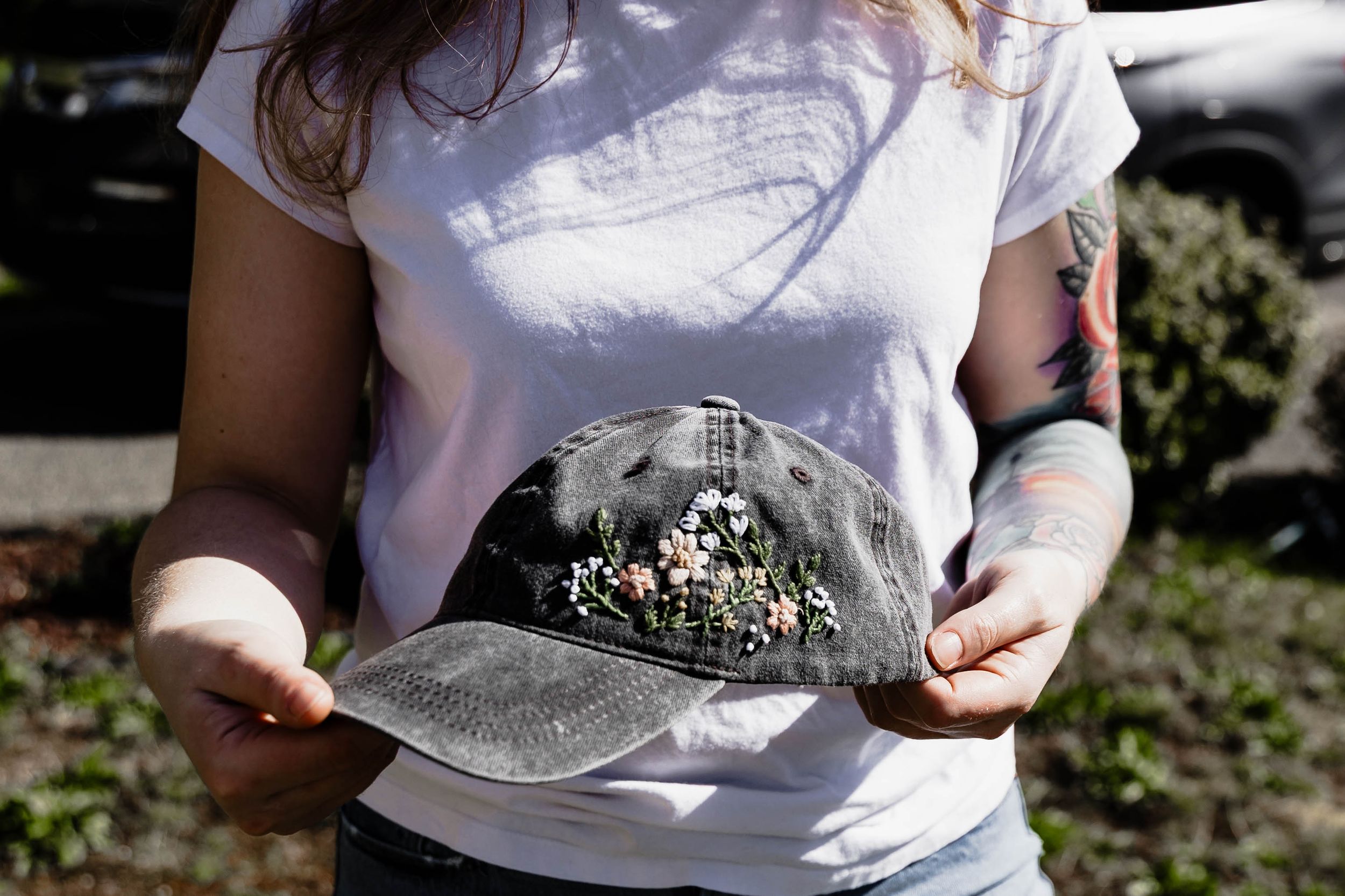 Someone holds a gray distressed baseball cap with an embroidered design while wearing a white t-shirt outdoors.