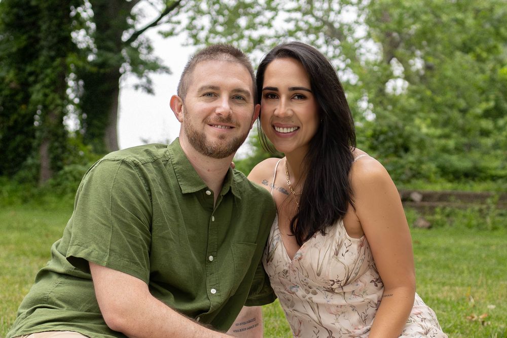 A couple sits together on grass in an outdoor setting with green trees in the background.