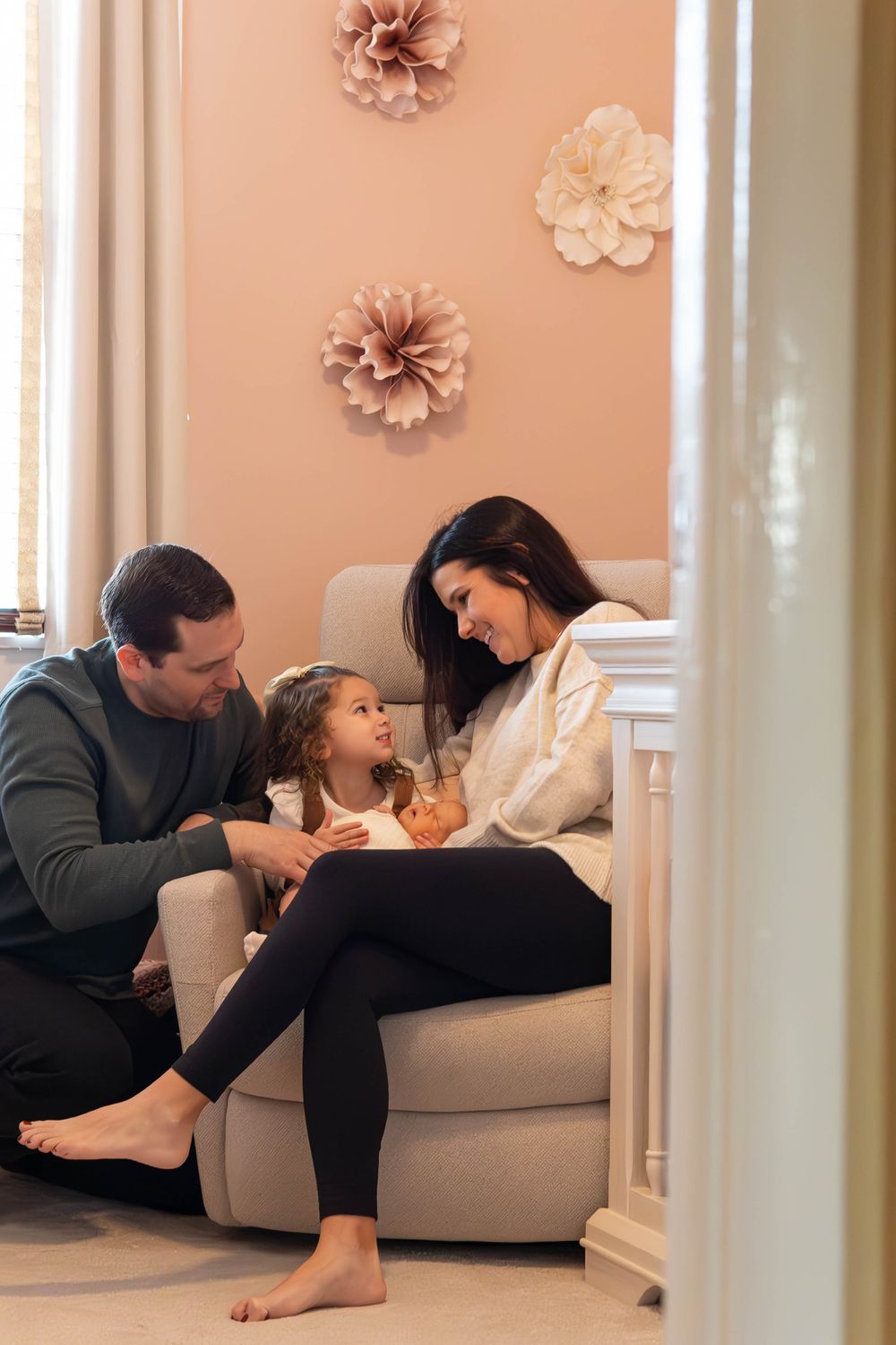 A family relaxes together on a couch in a living room with soft peach walls decorated with paper flowers.