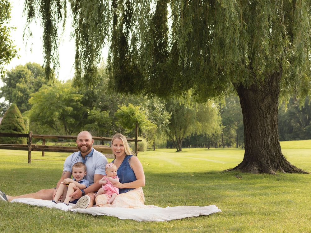 Family enjoying a summer picnic together on a blanket under willow trees in a peaceful park setting.