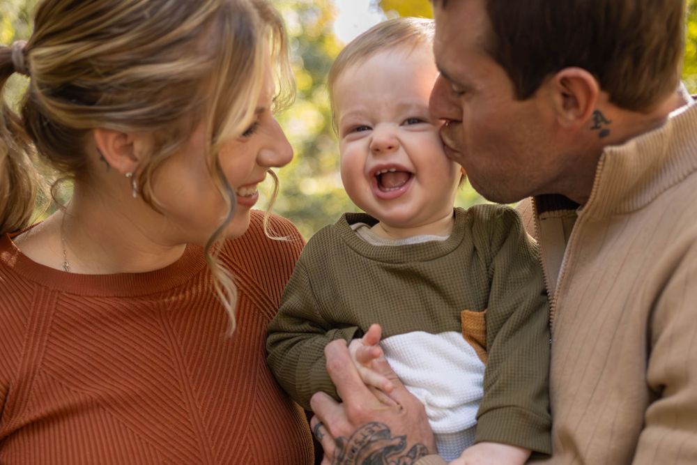 A joyful baby laughs while being held by parents outdoors in warm autumn lighting.