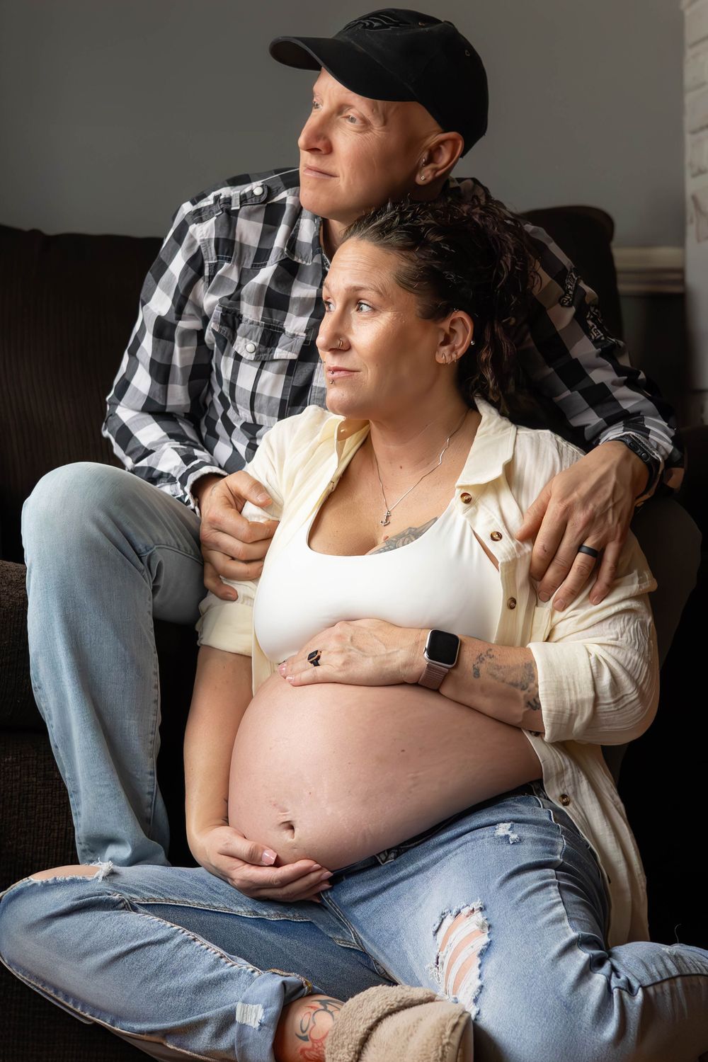 A pregnant person in a white top and jeans sits with their partner in a plaid shirt in a series of intimate photos.