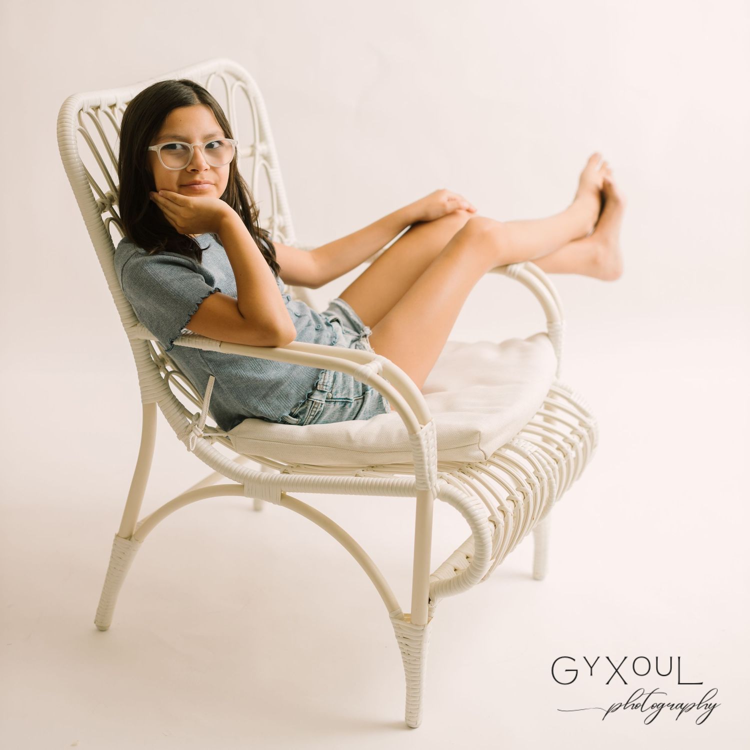 A relaxed portrait in a white wicker chair showing casual denim style against neutral backdrop.