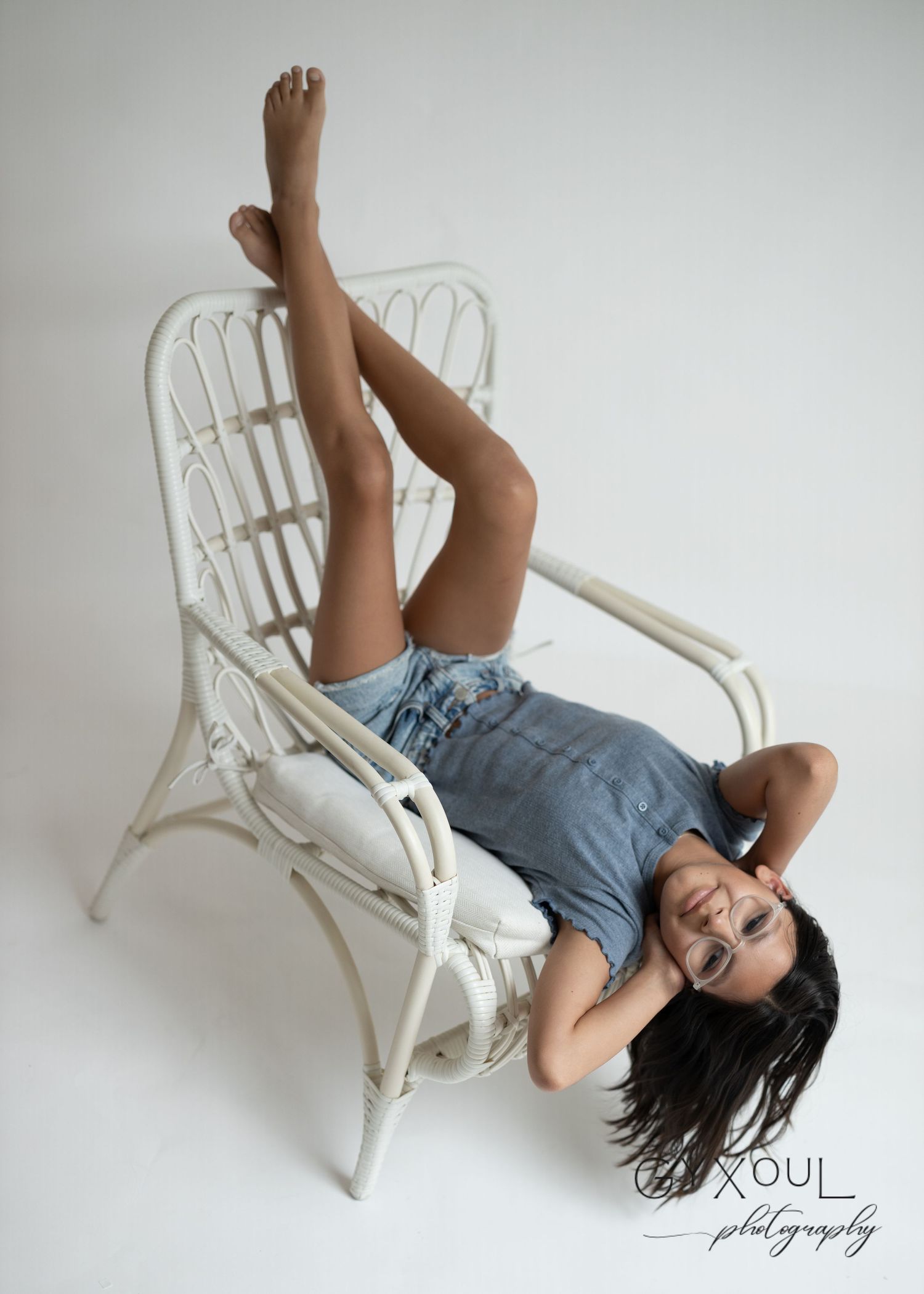 Creative portrait on white wicker chair with playful upside-down pose in casual denim outfit.