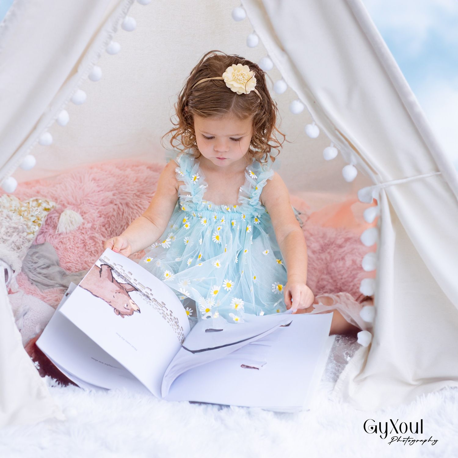 Small child in blue floral dress reading a book inside a white teepee tent with pink pillows.
