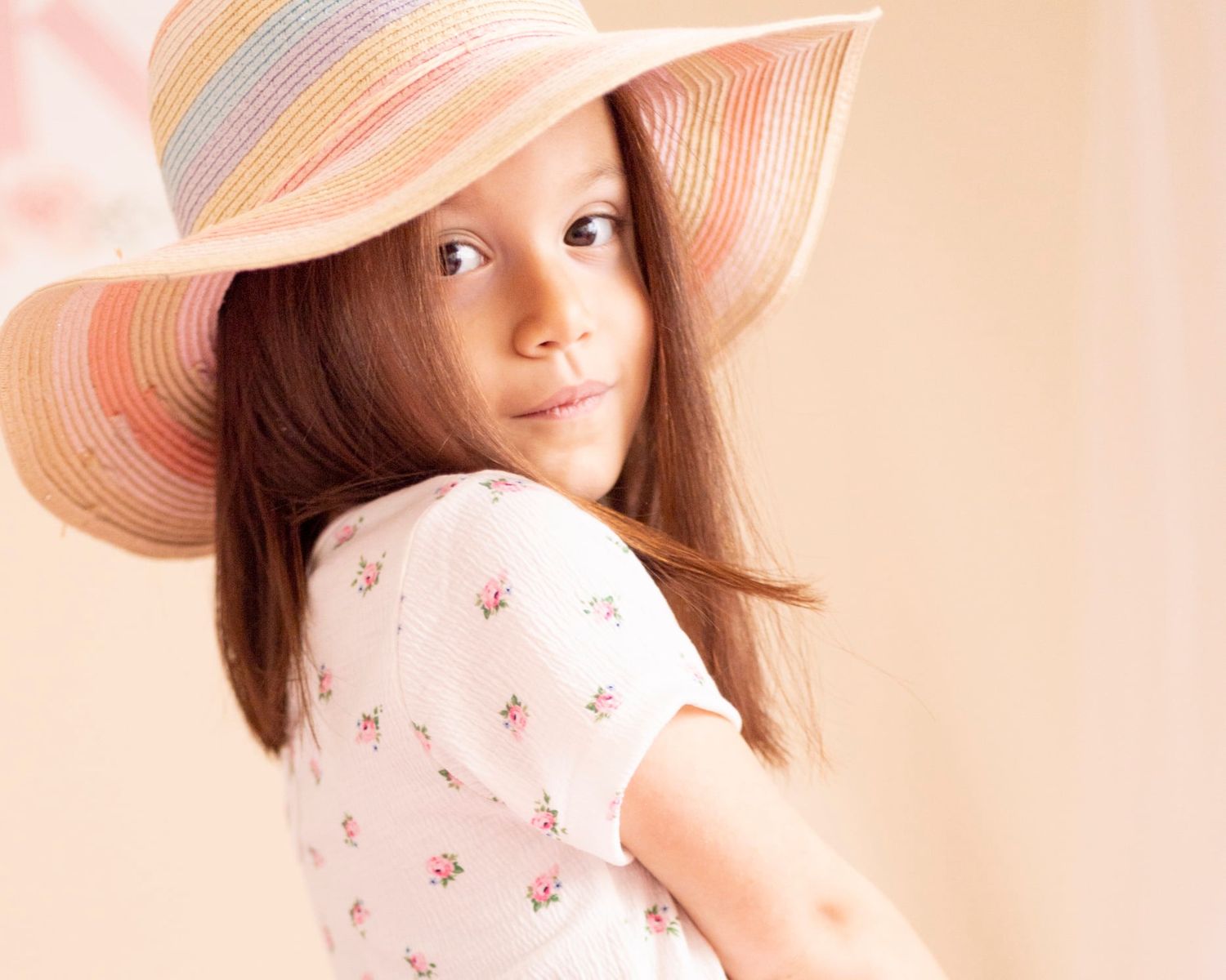 Portrait of a person wearing a pink floral dress and large sun hat against a peachy background.