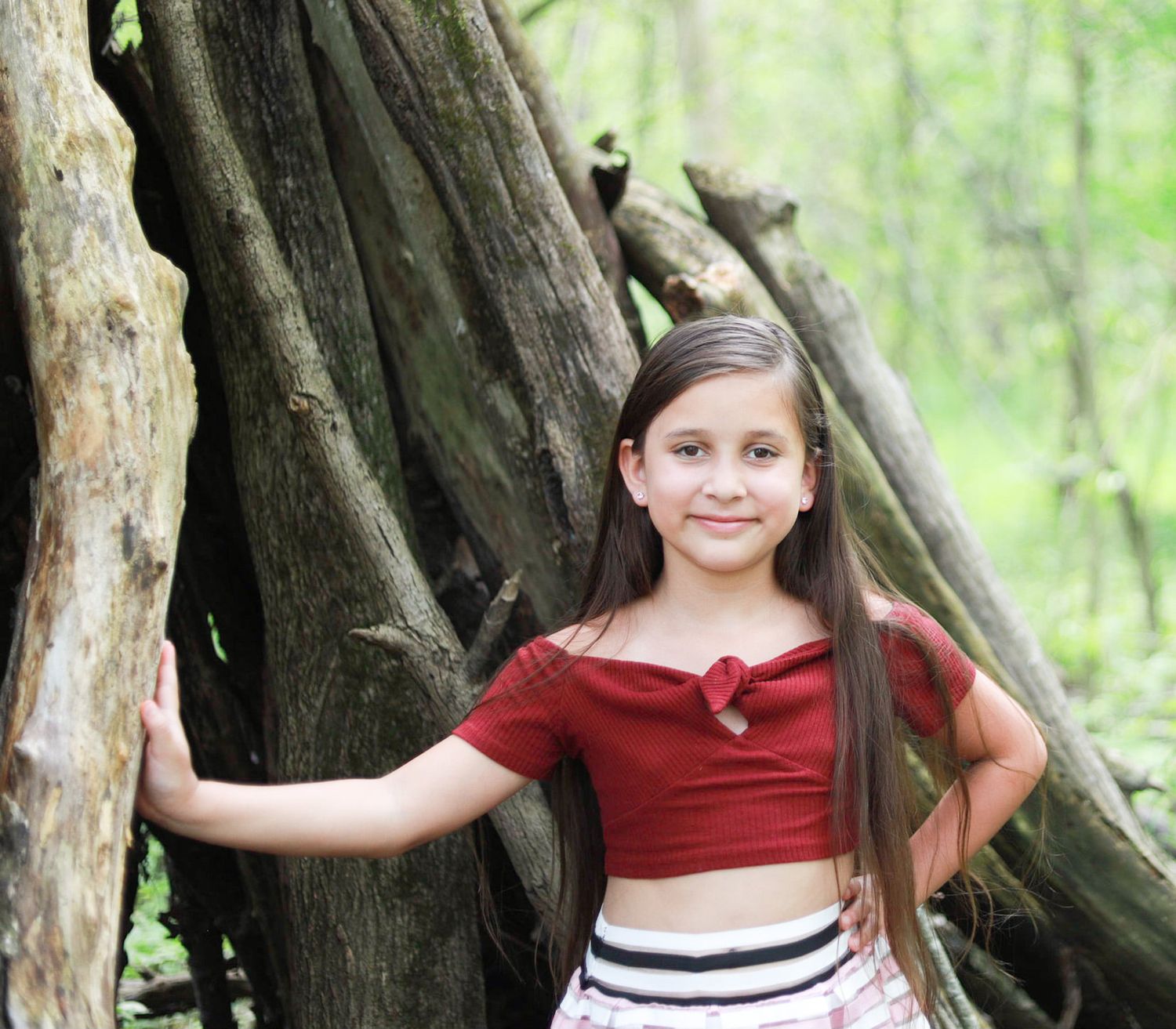 A girl in a red crop top poses by a large tree trunk in a forest setting.