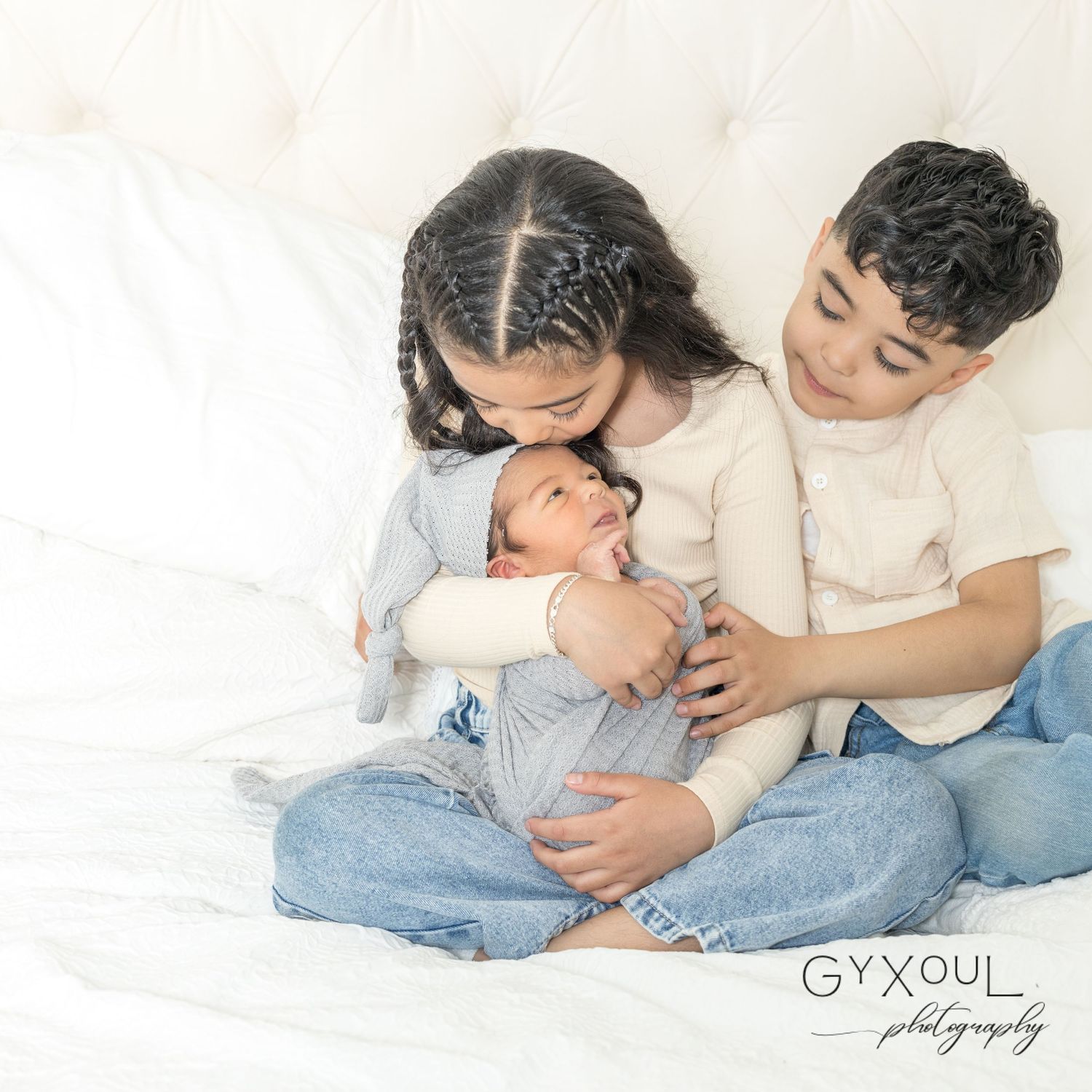 Intimate family moment showing siblings bonding with newborn baby on white bedding.