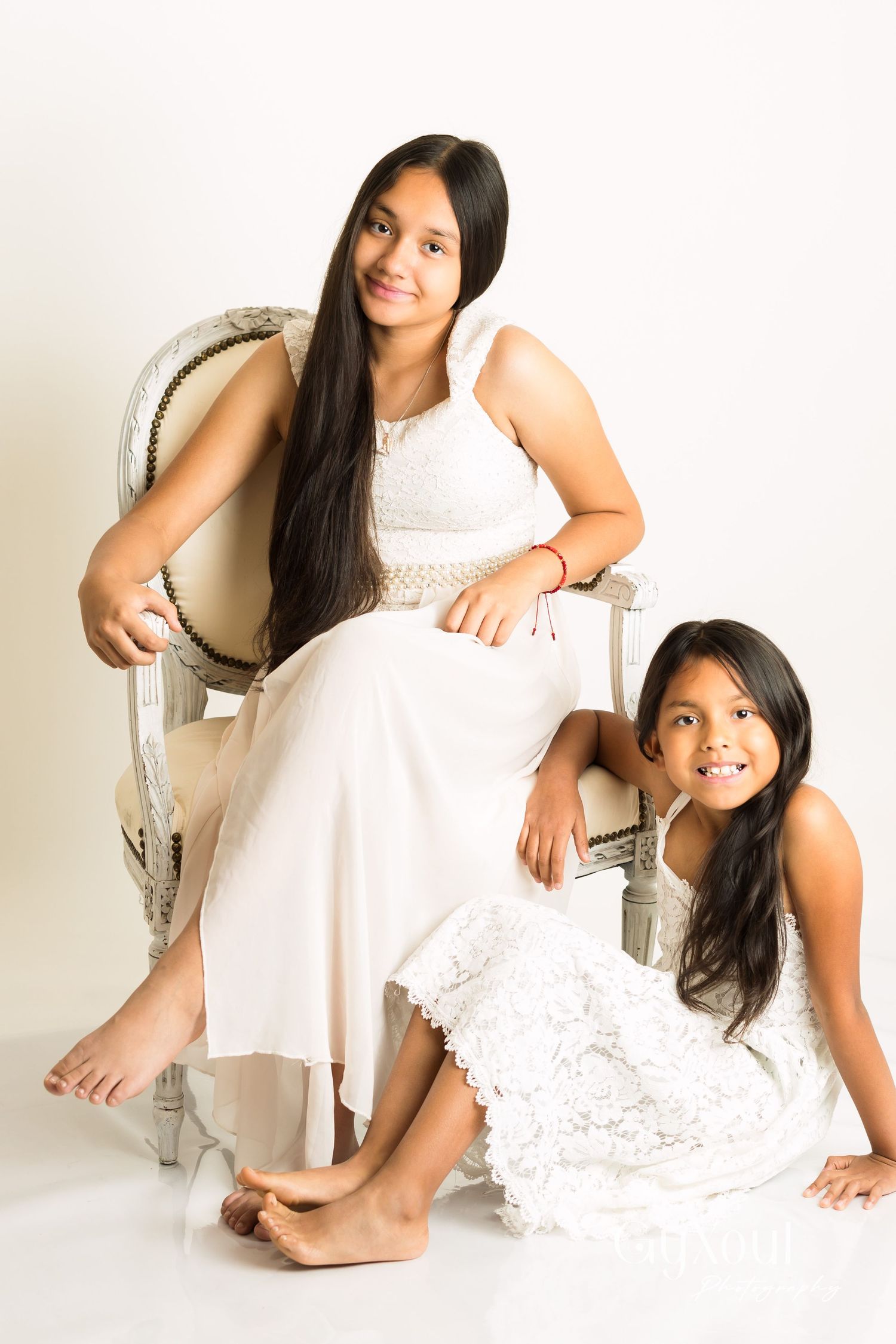A tasteful portrait of two people in white dresses sitting on an elegant white chair against a light background.