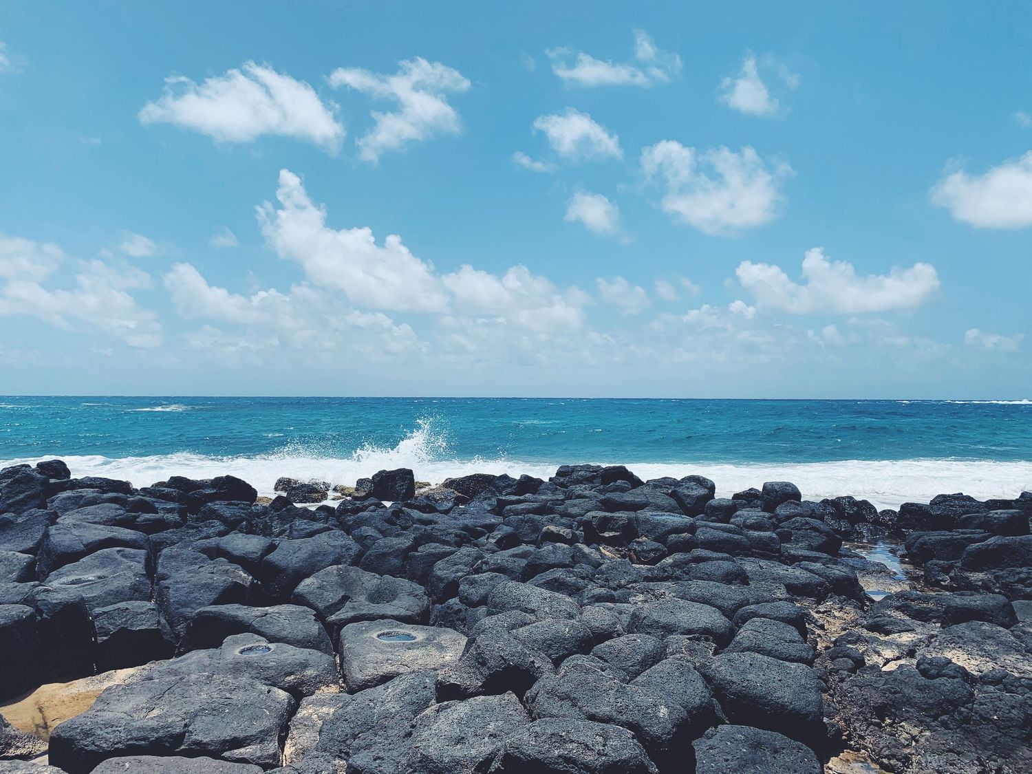 Rocky volcanic coastline with crashing ocean waves under bright blue sky with white clouds.