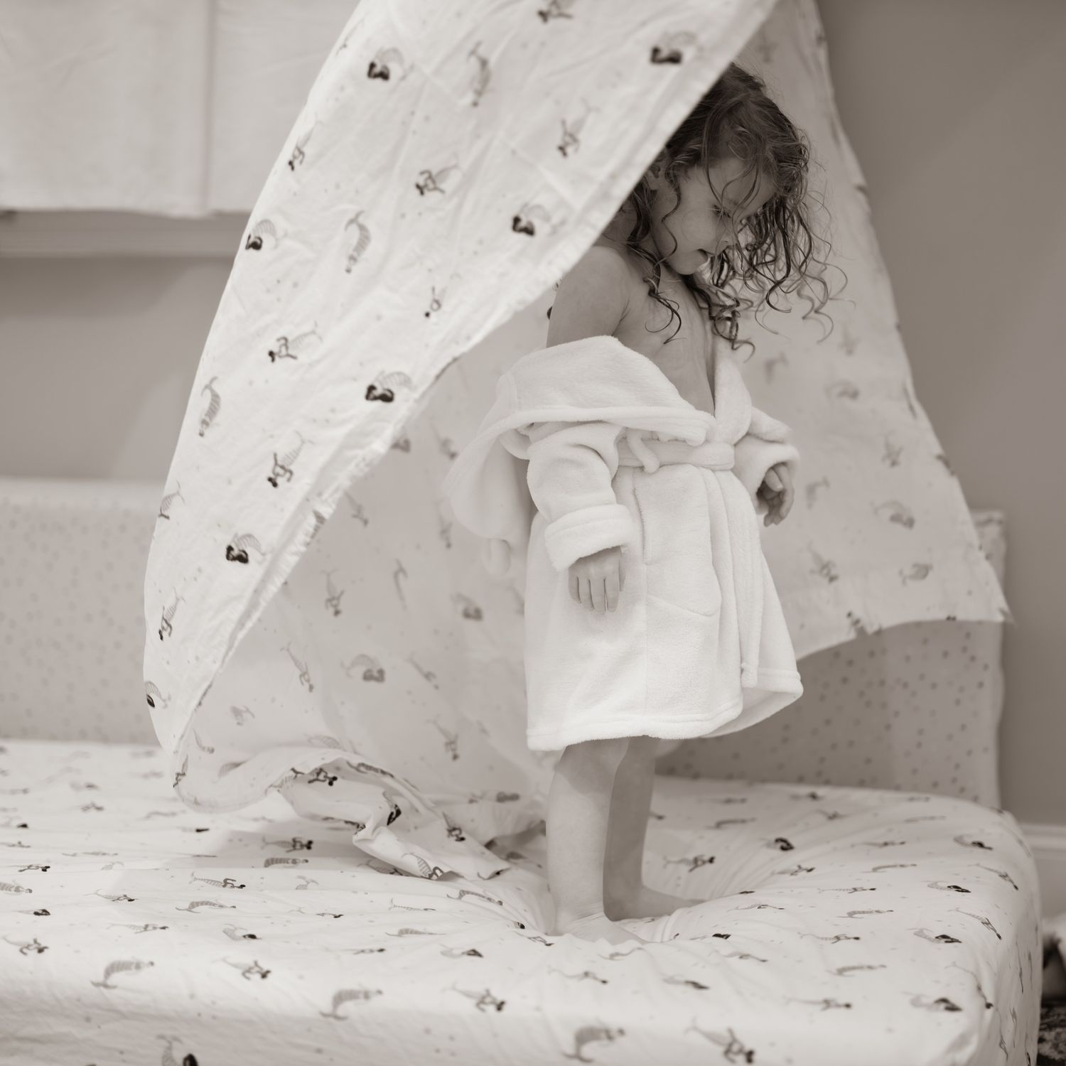 Black and white photo of child in white dress playing on patterned bedding.