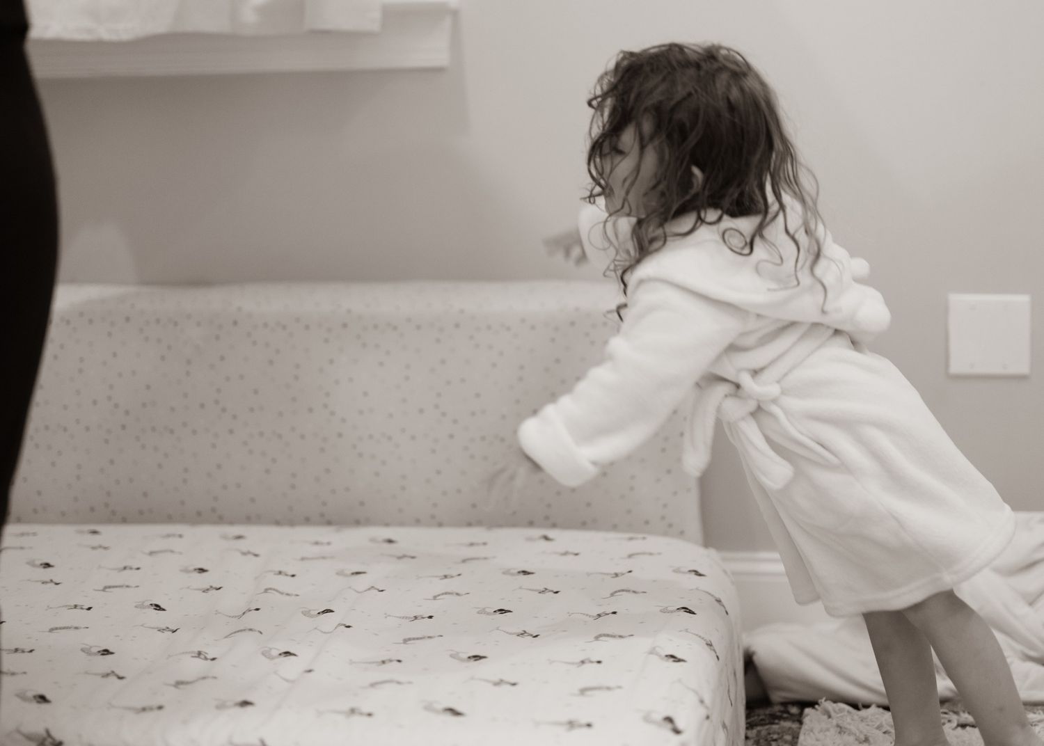 Black and white image of child in white robe climbing on bed.