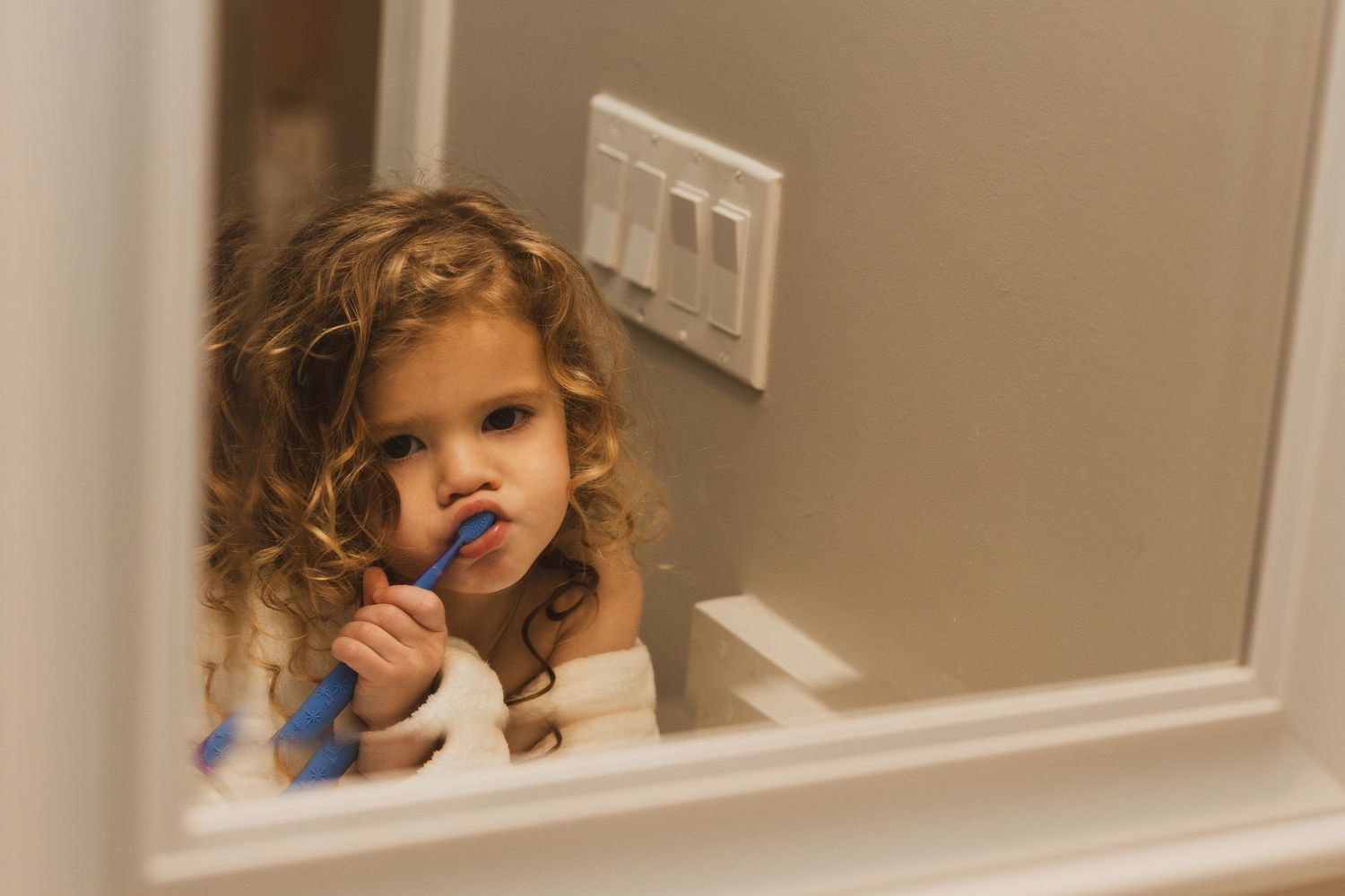 Young child with curly hair gazes into bathroom mirror while brushing teeth.