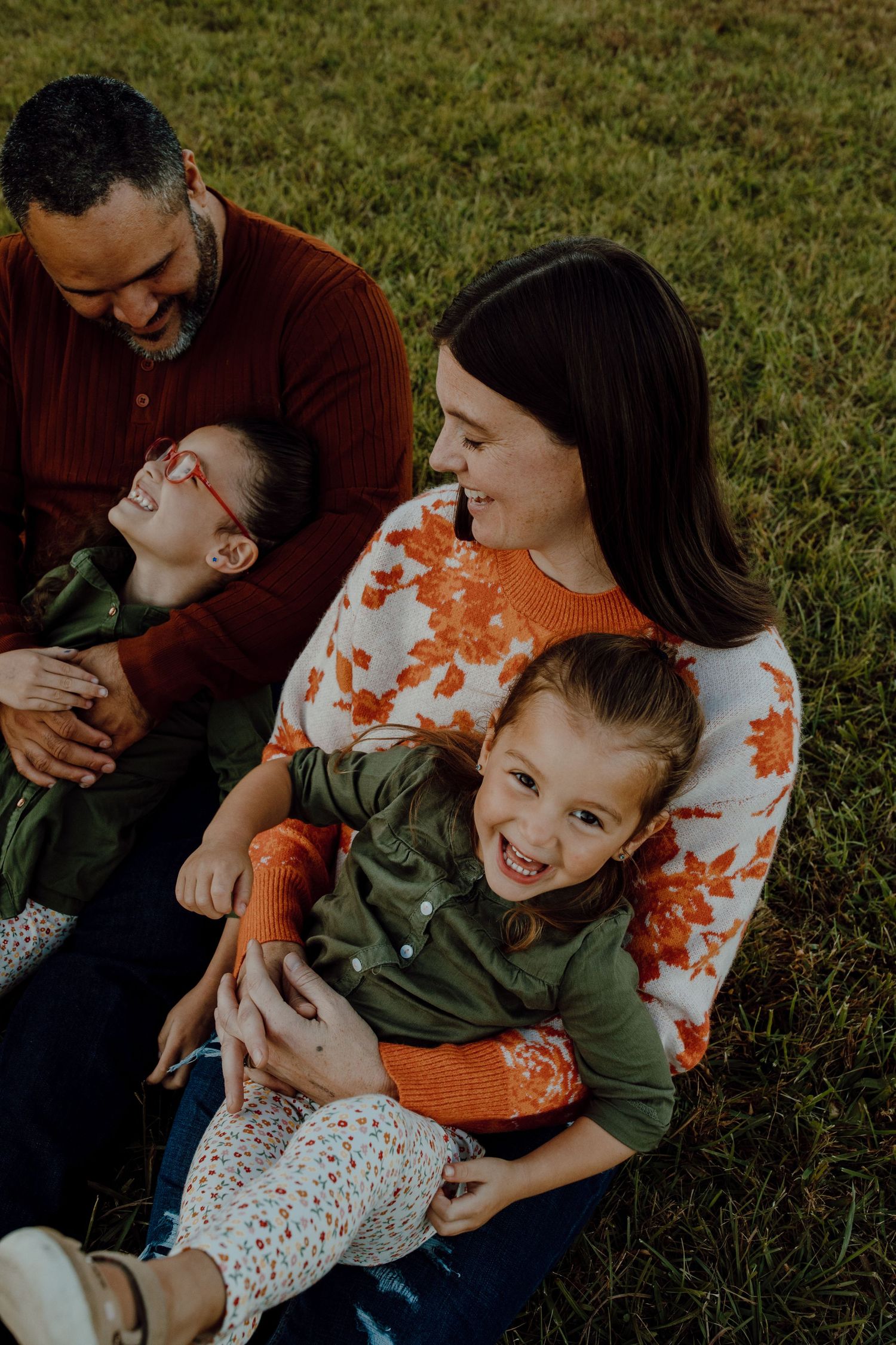 A family shares joyful laughter while sitting together on grass during an outdoor photo session.