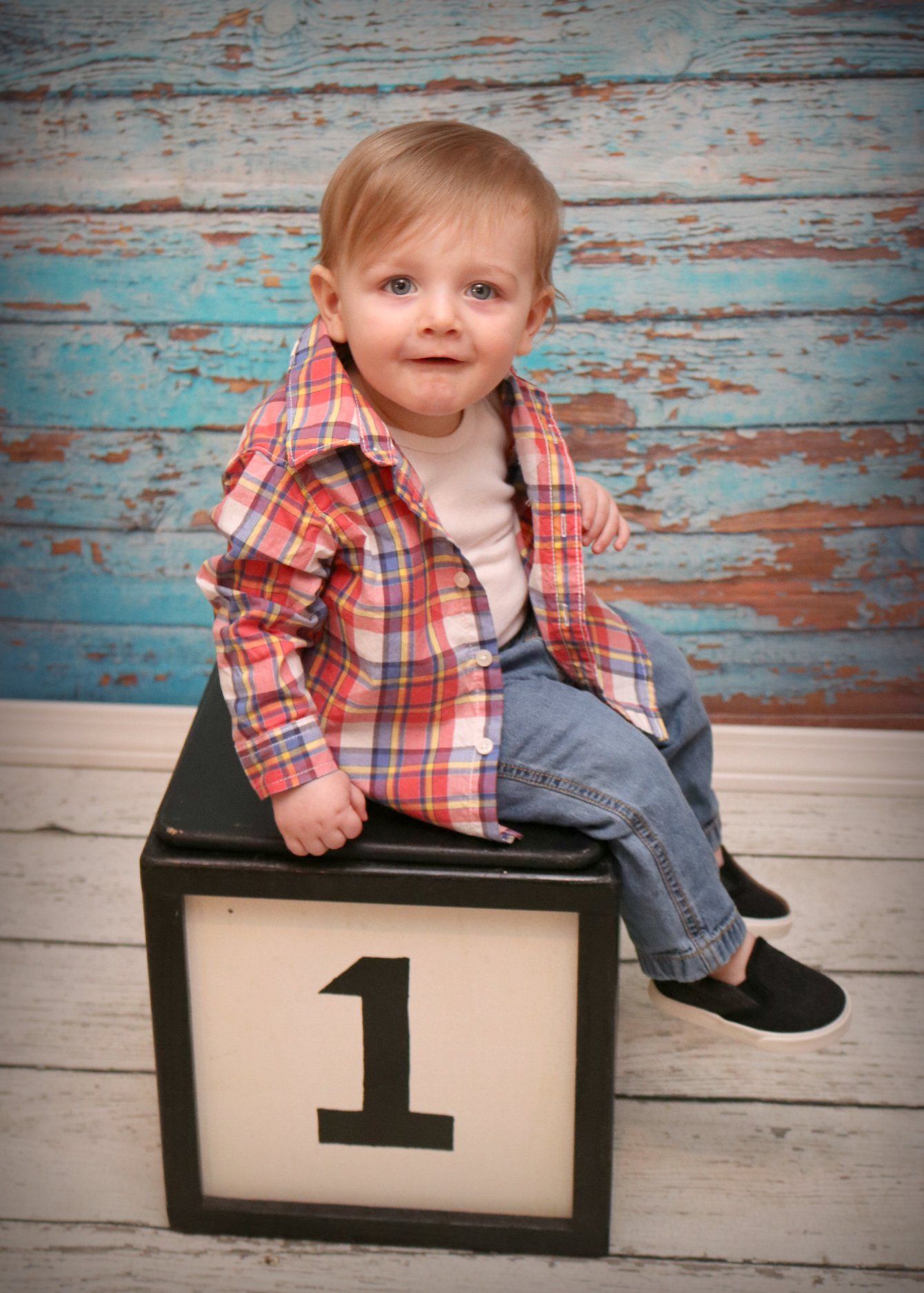 Baby in plaid shirt sits on wooden number one block against weathered blue backdrop.