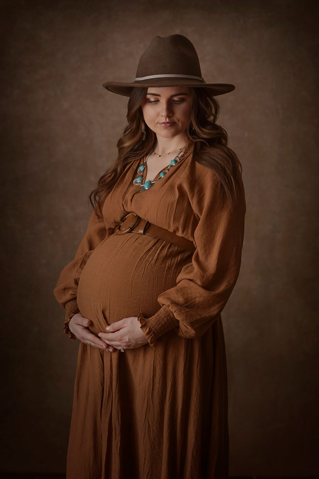 Maternity photo featuring brown dress and western hat with turquoise jewelry against moody brown backdrop. Blackfoot maternity photographer