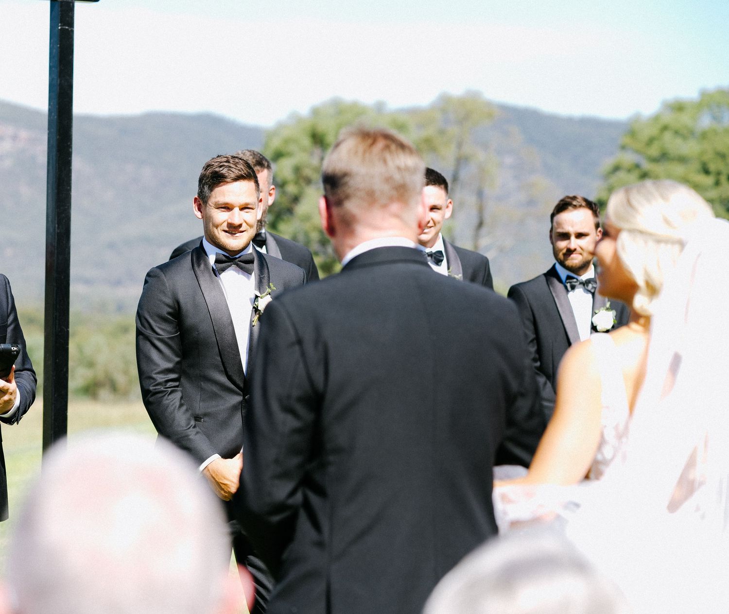 Wedding ceremony attendees gather outdoors with mountains visible in background.