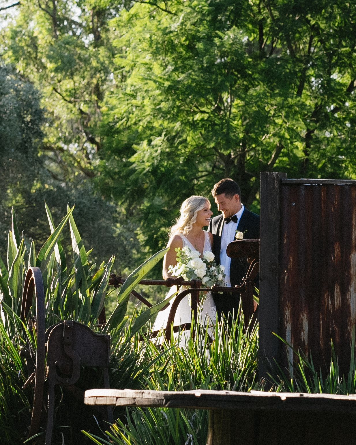 Couple shares intimate moment on wooden deck surrounded by tall grass and greenery.