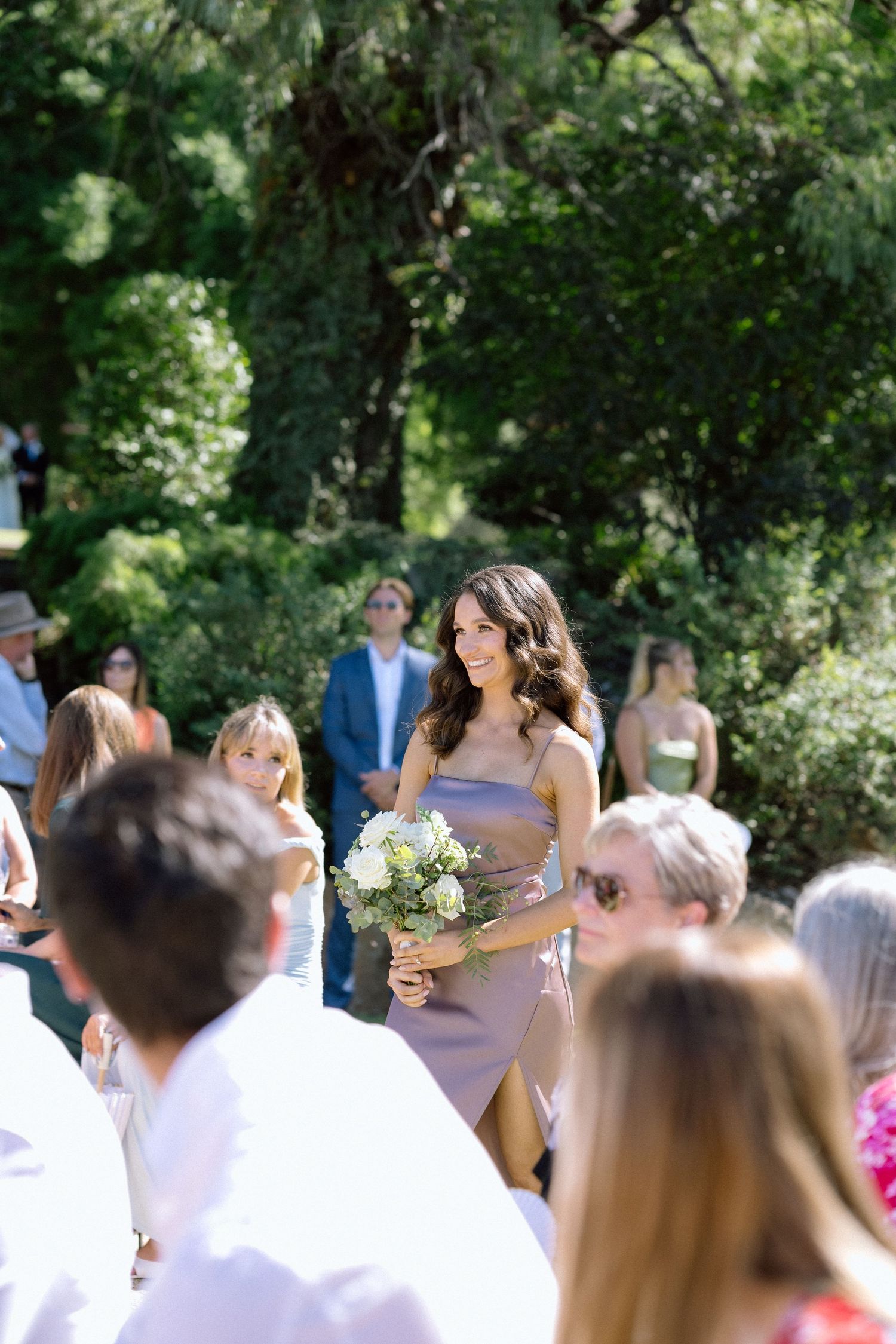A bridesmaid in a lavender dress walks down an outdoor aisle carrying a white bouquet during a garden ceremony.