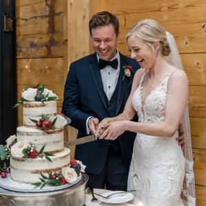 Newlywed couple cutting their elegant white wedding cake decorated with burgundy flowers and greenery together.