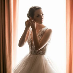 Silhouetted figure in a flowing white wedding gown with lace details puts on earrings against backlit peach curtains.