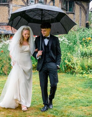 A couple shares an umbrella while walking through a garden on a rainy wedding day, surrounded by blooming flowers.