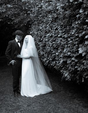 A wedding couple stands together beside a green garden hedge in black and white photography.