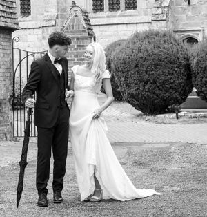 Black and white photo of a bride and groom outside a historic church building with an umbrella and rounded shrub.