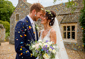 Happy newlyweds embrace while being showered with colorful confetti outside a stone country house as bride holds floral bouquet.
