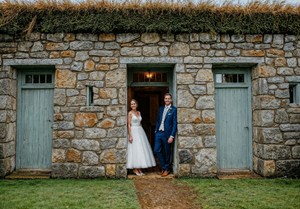 A vintage stone building with light blue wooden doors and a couple standing in the illuminated doorway at dusk.