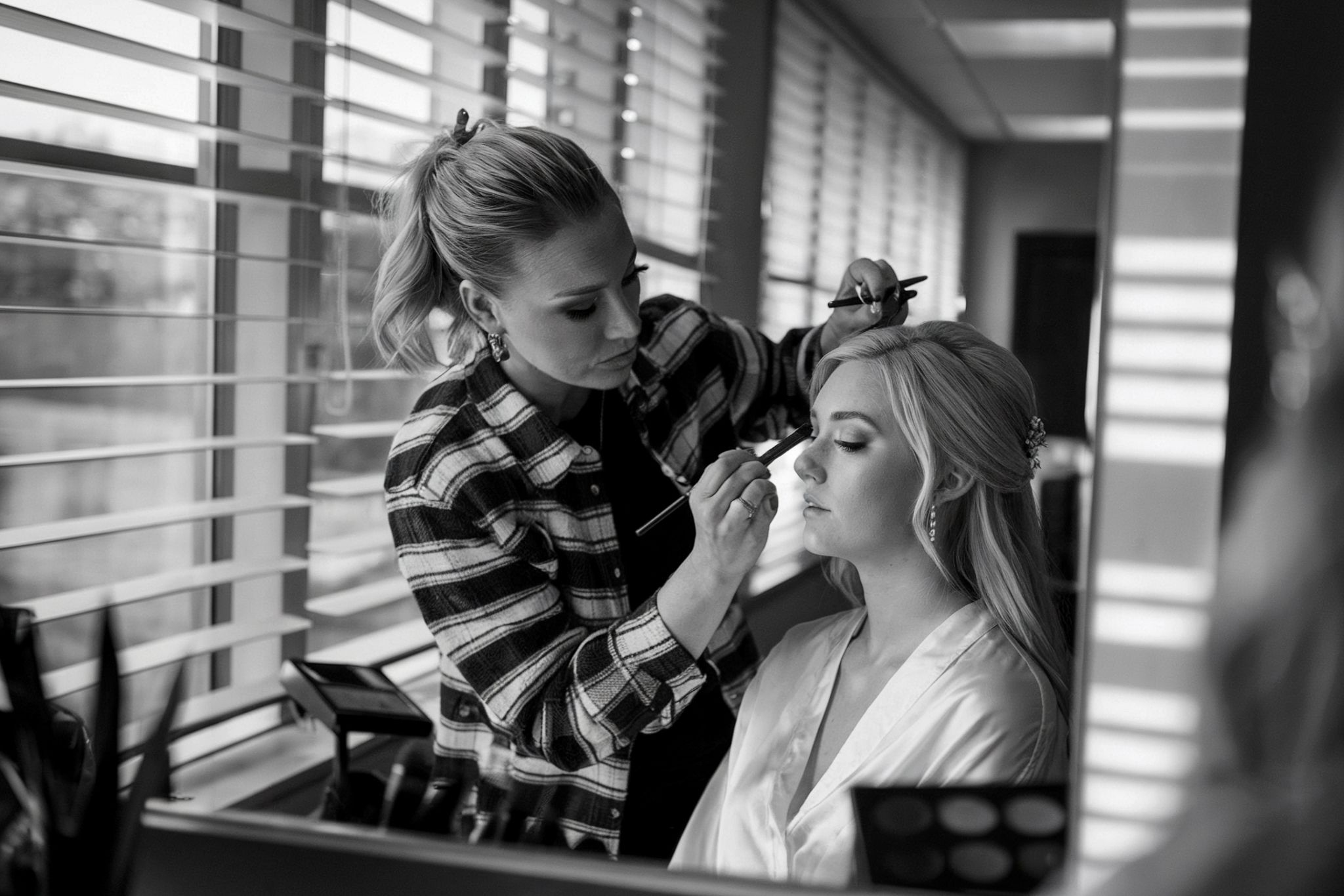 Black and white image of bride getting makeup applied near window with natural light.