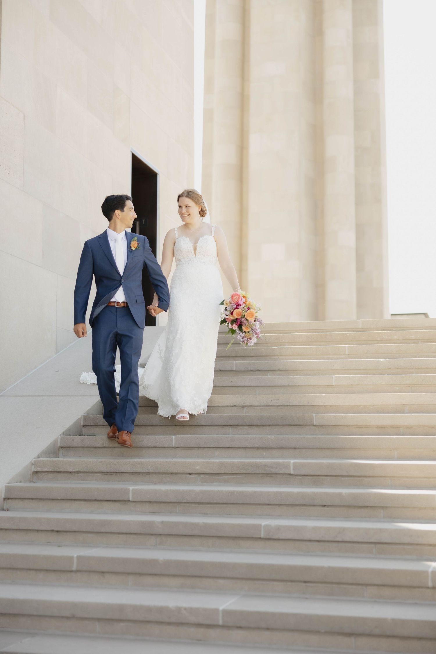 A couple in wedding attire walks down wide stone steps at an elegant venue with white columns in the background.