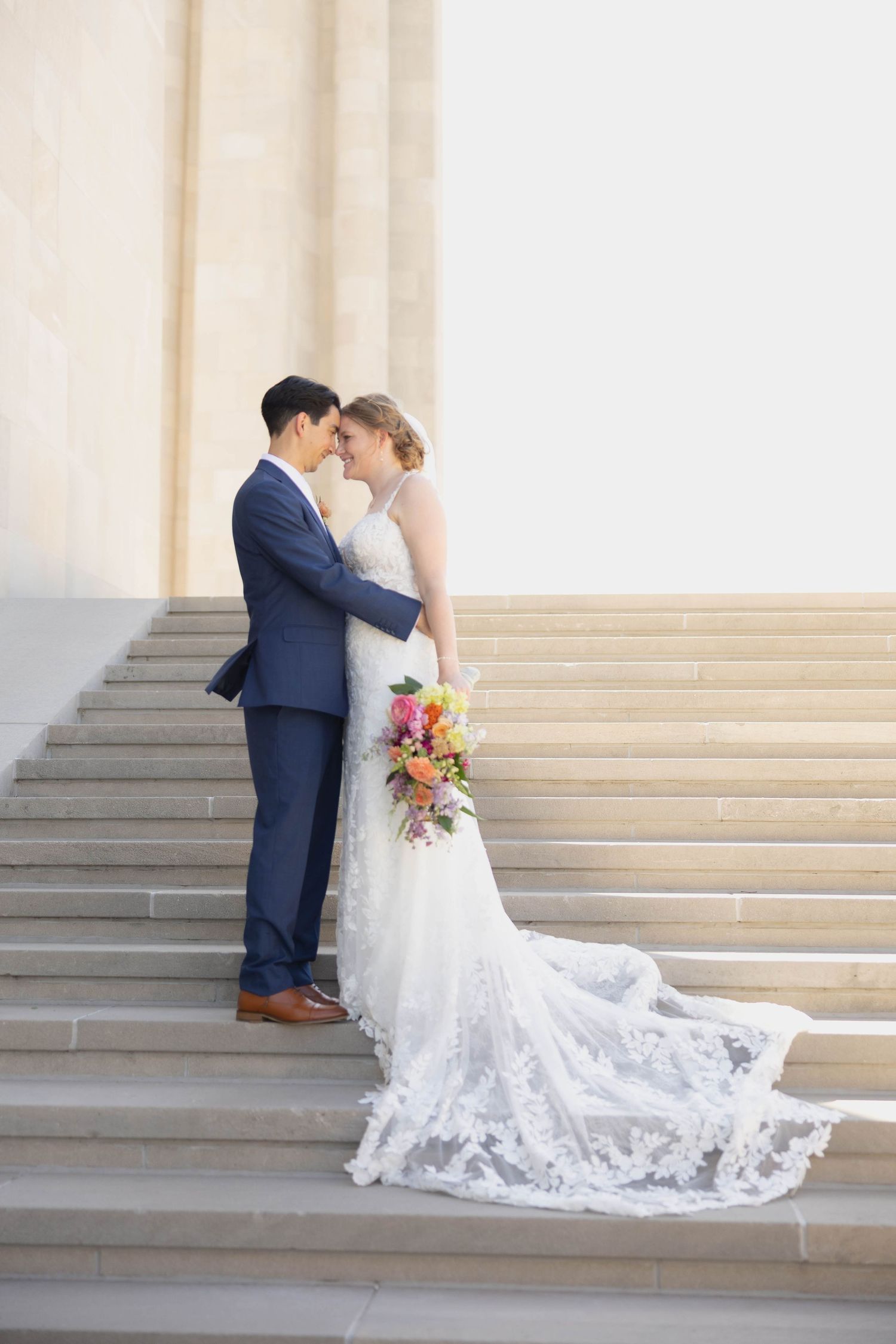 A romantic wedding couple embraces on stone steps with bride's lace dress trailing elegantly behind them.