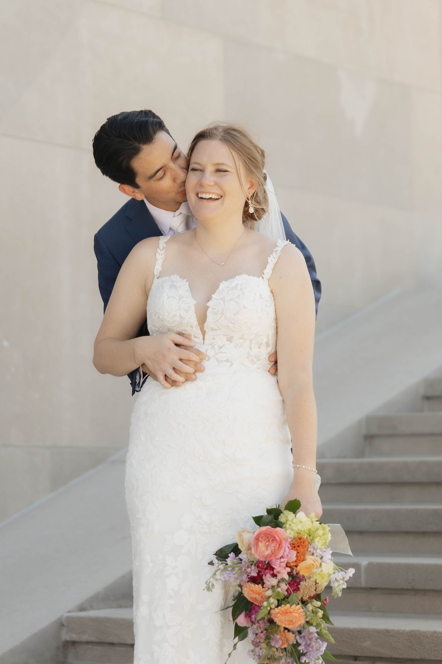 A romantic wedding couple embraces on stone steps with a colorful pink and peach floral bouquet.