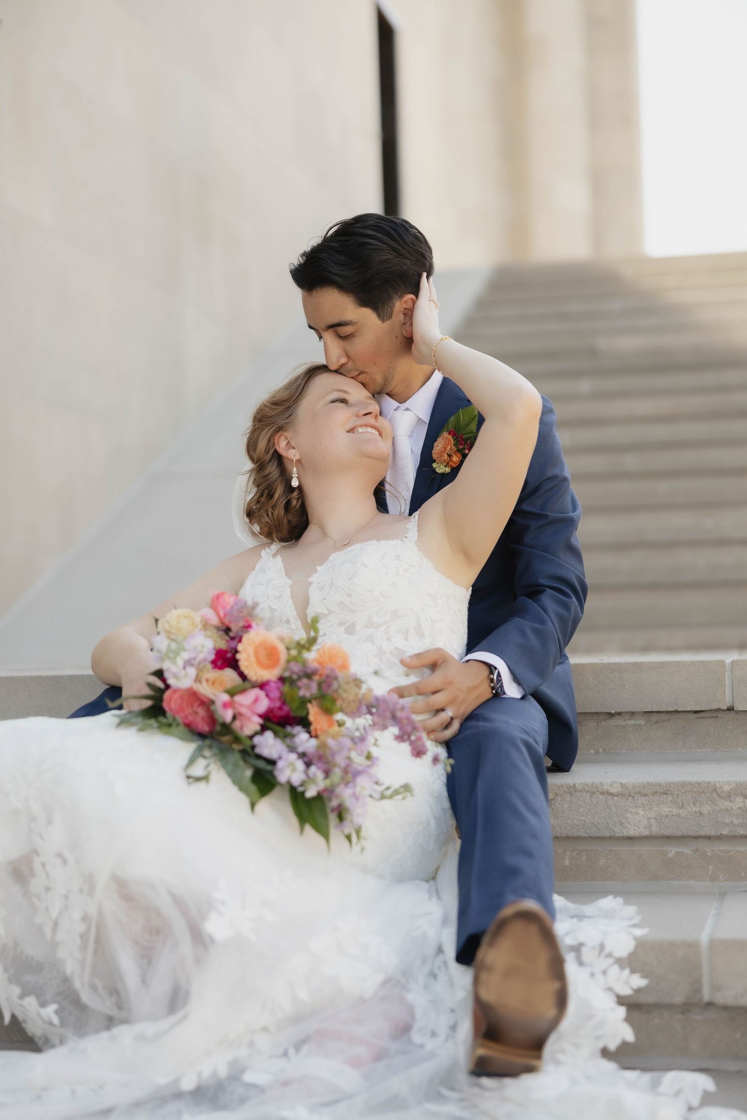 Newlyweds share an intimate moment on marble steps with colorful bouquet and flowing white lace gown.