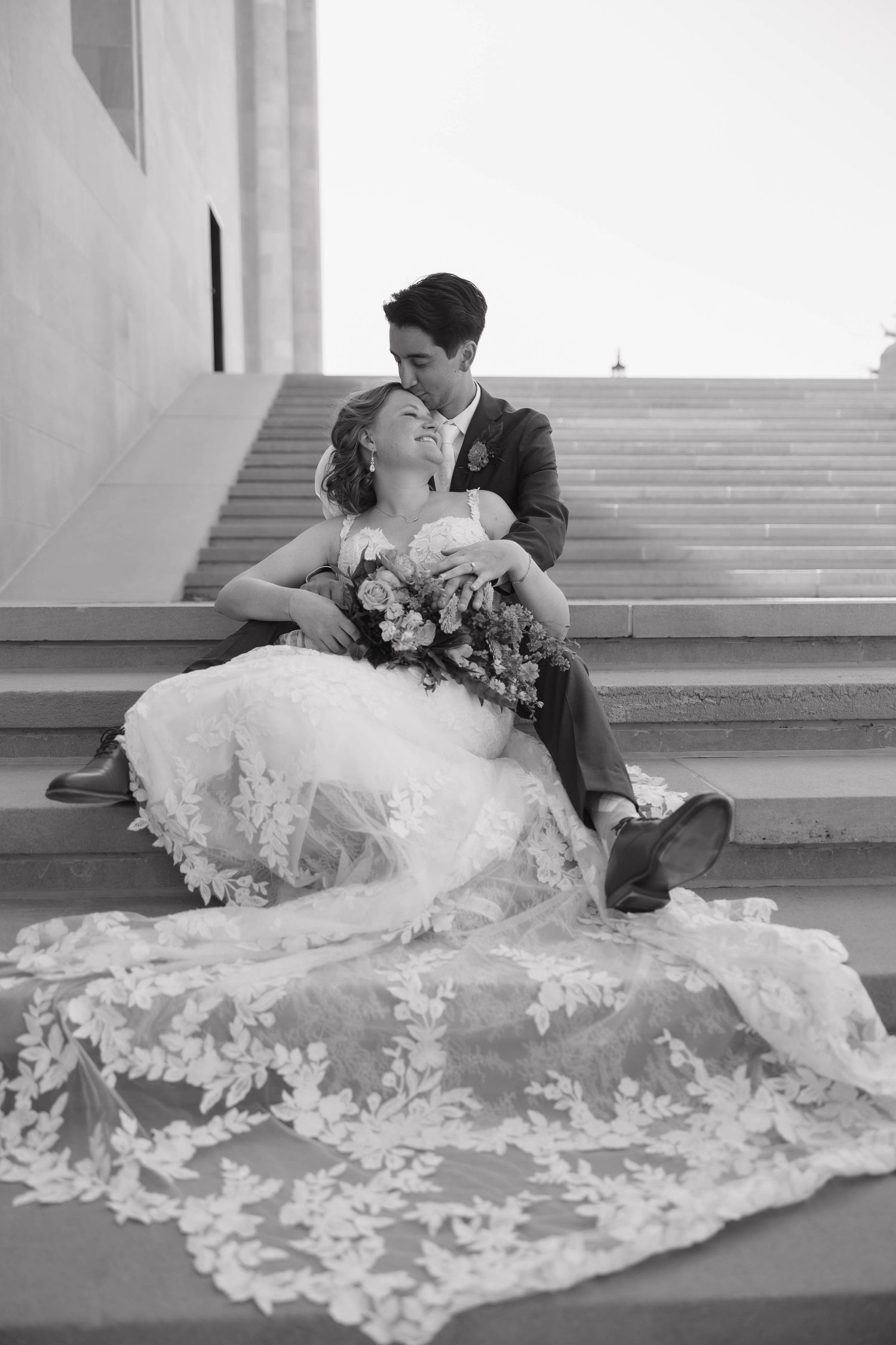 Black and white wedding photo captures couple's tender moment on grand staircase with dramatic lace train.