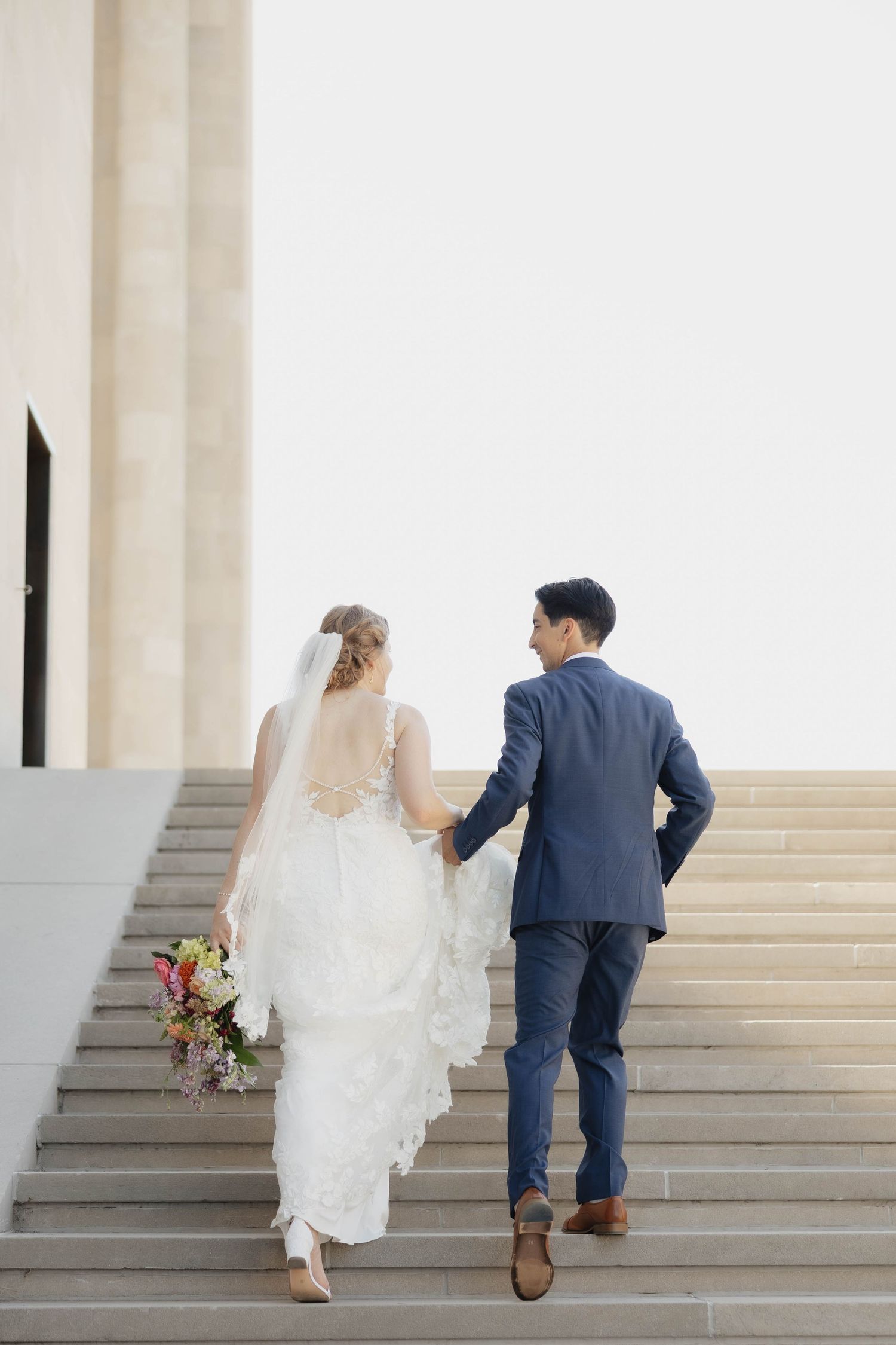 A newlywed couple ascends stone stairs together, with the bride in a flowing white dress holding a floral bouquet.