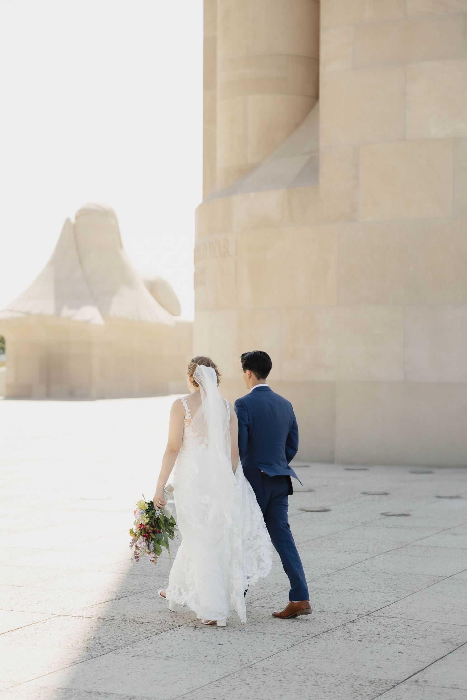 Newlyweds walk together on a light stone terrace with beautiful architectural columns in the background.