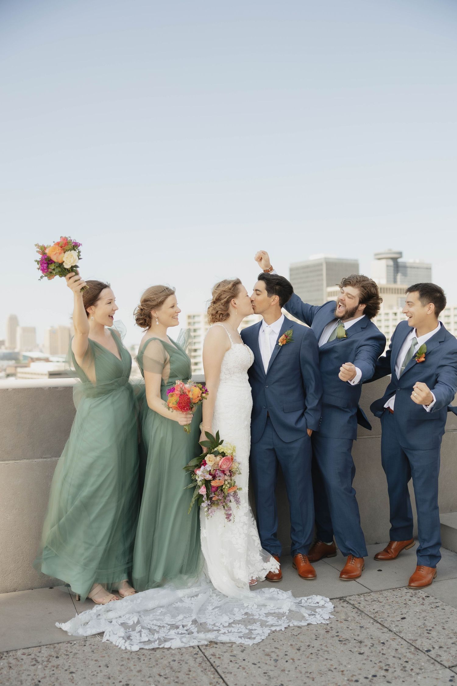 Wedding party in navy suits and sage green dresses pose playfully against a city skyline.