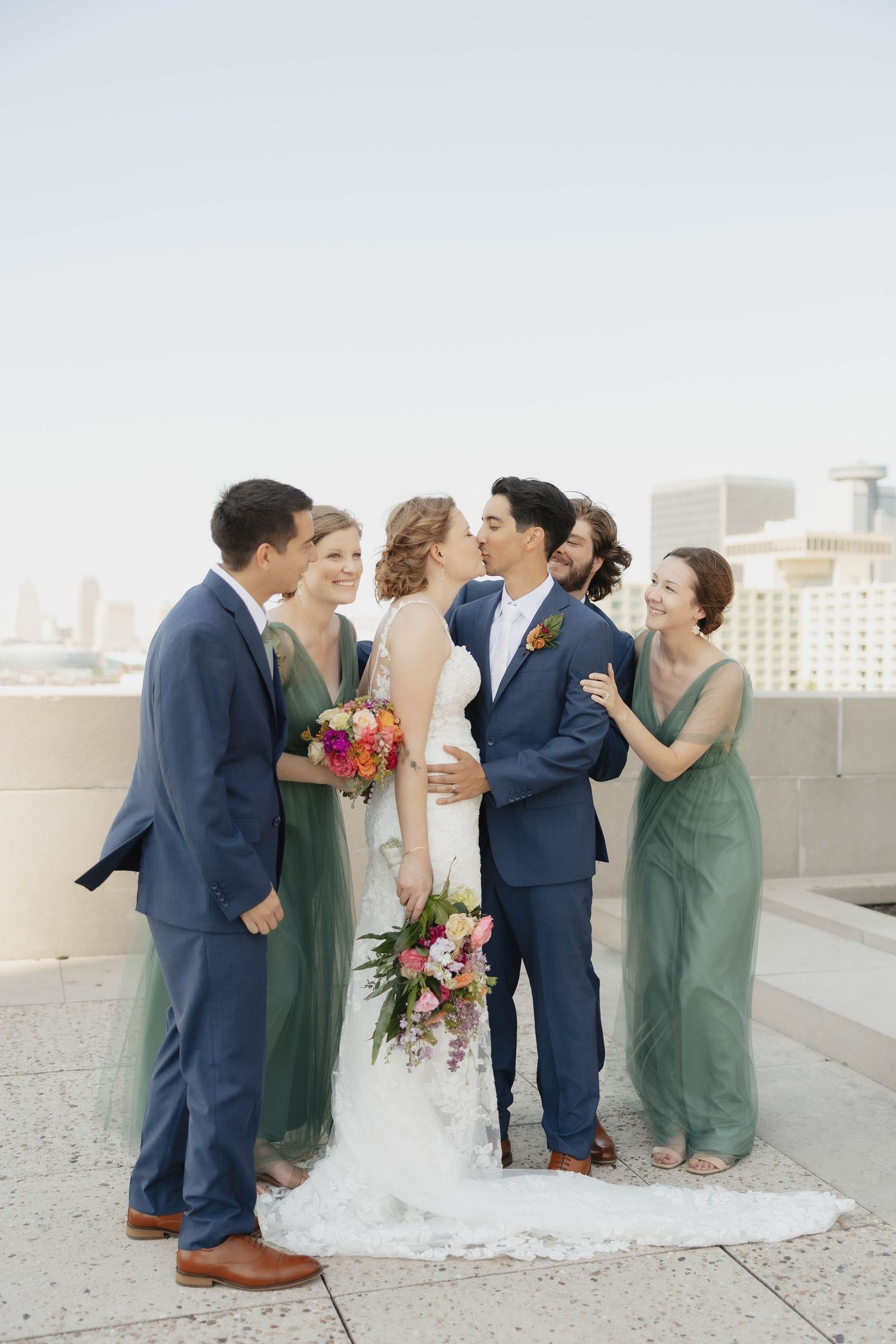 Wedding party members in navy suits and sage dresses share candid laughs on a rooftop venue.